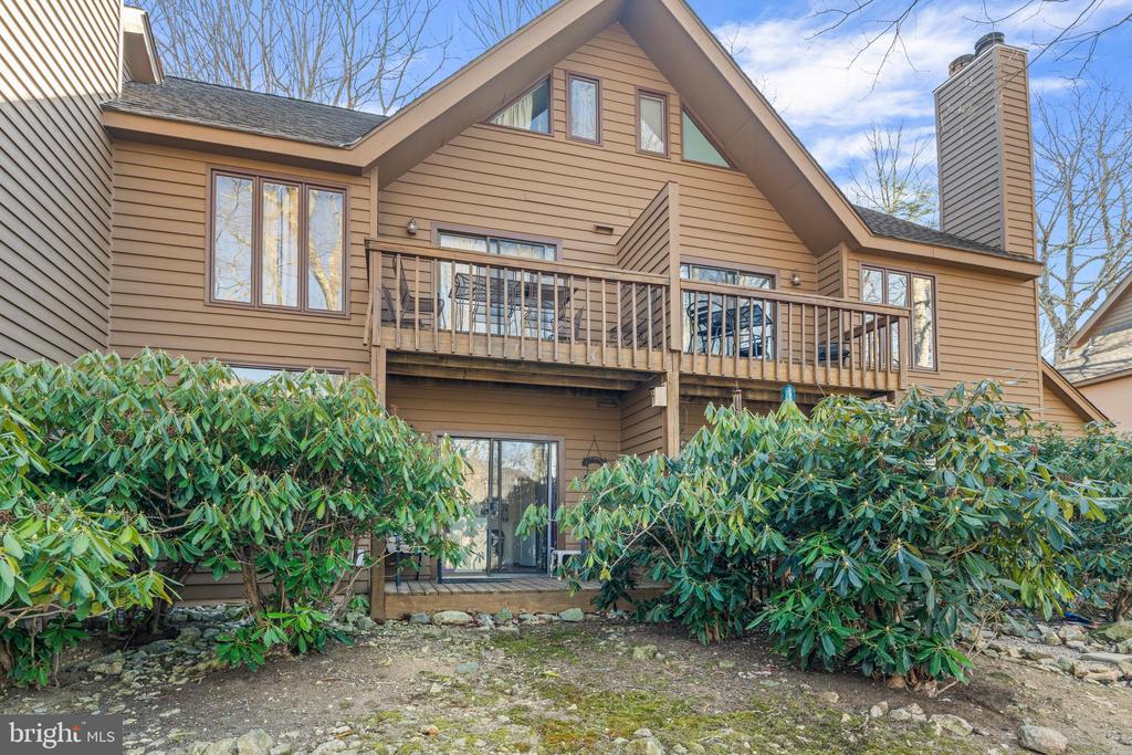 House rear with elevated wooden deck, steep gabled roof, and dense shrubs.