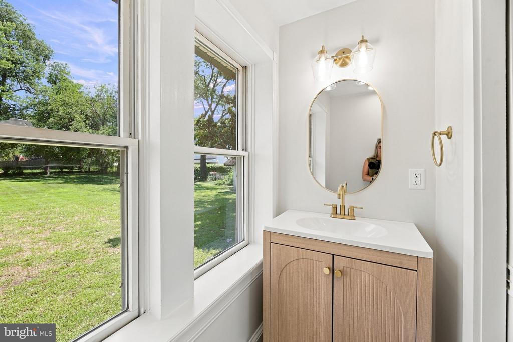Bathroom vanity with a light wood cabinet and gold fixtures beside large windows overlooking a green lawn.