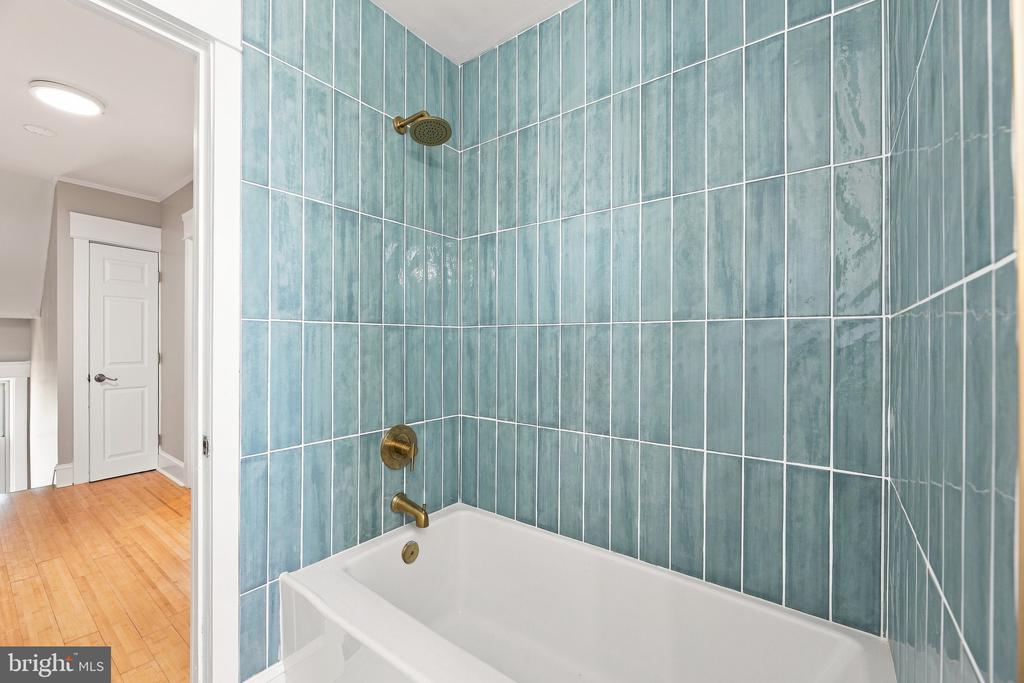 Bathroom with a white tub, blue-green tiled shower, and brass fixtures beside a hardwood hallway.