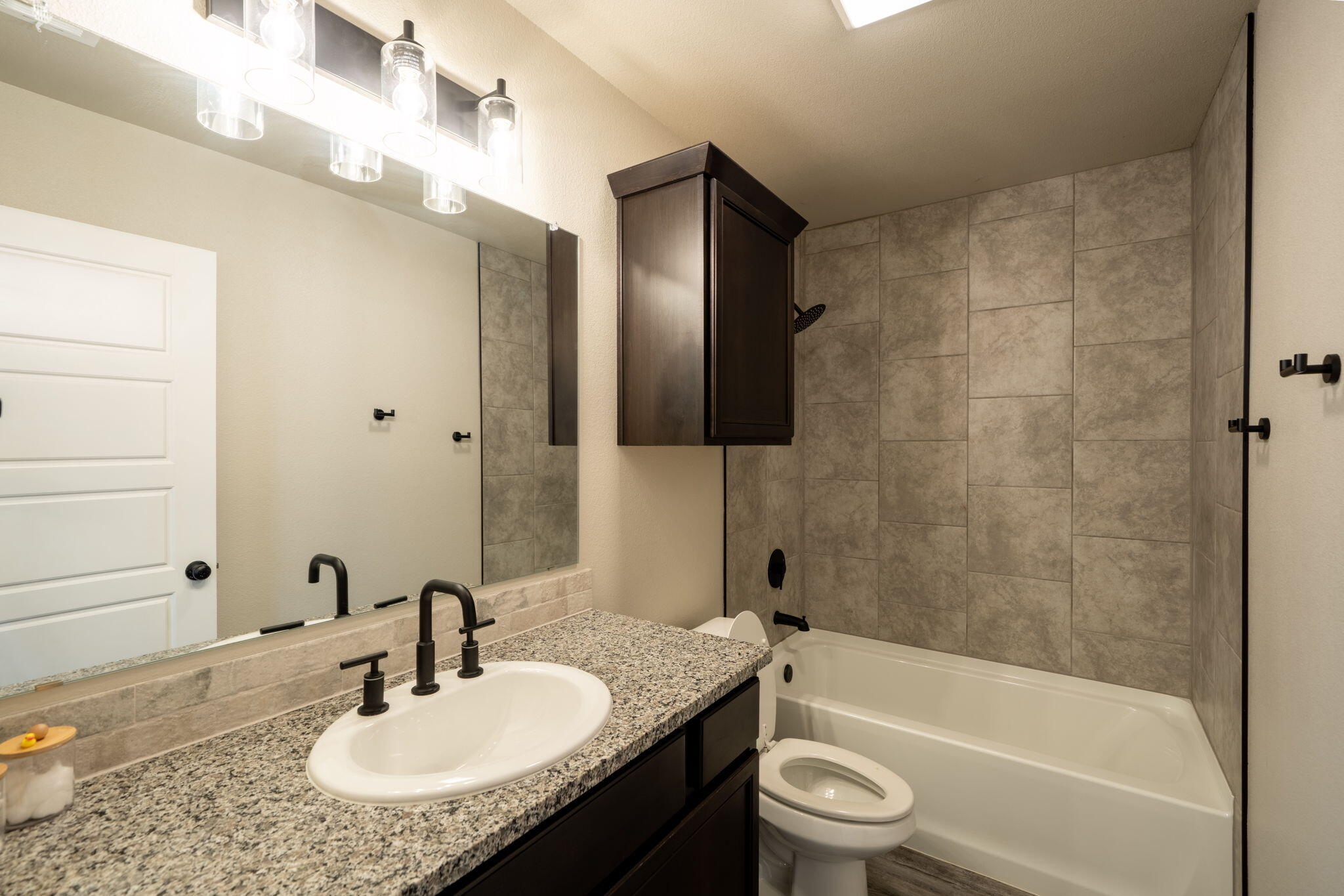 Bathroom with a granite vanity, dark cabinetry, and gray tile surrounding the tub and shower.