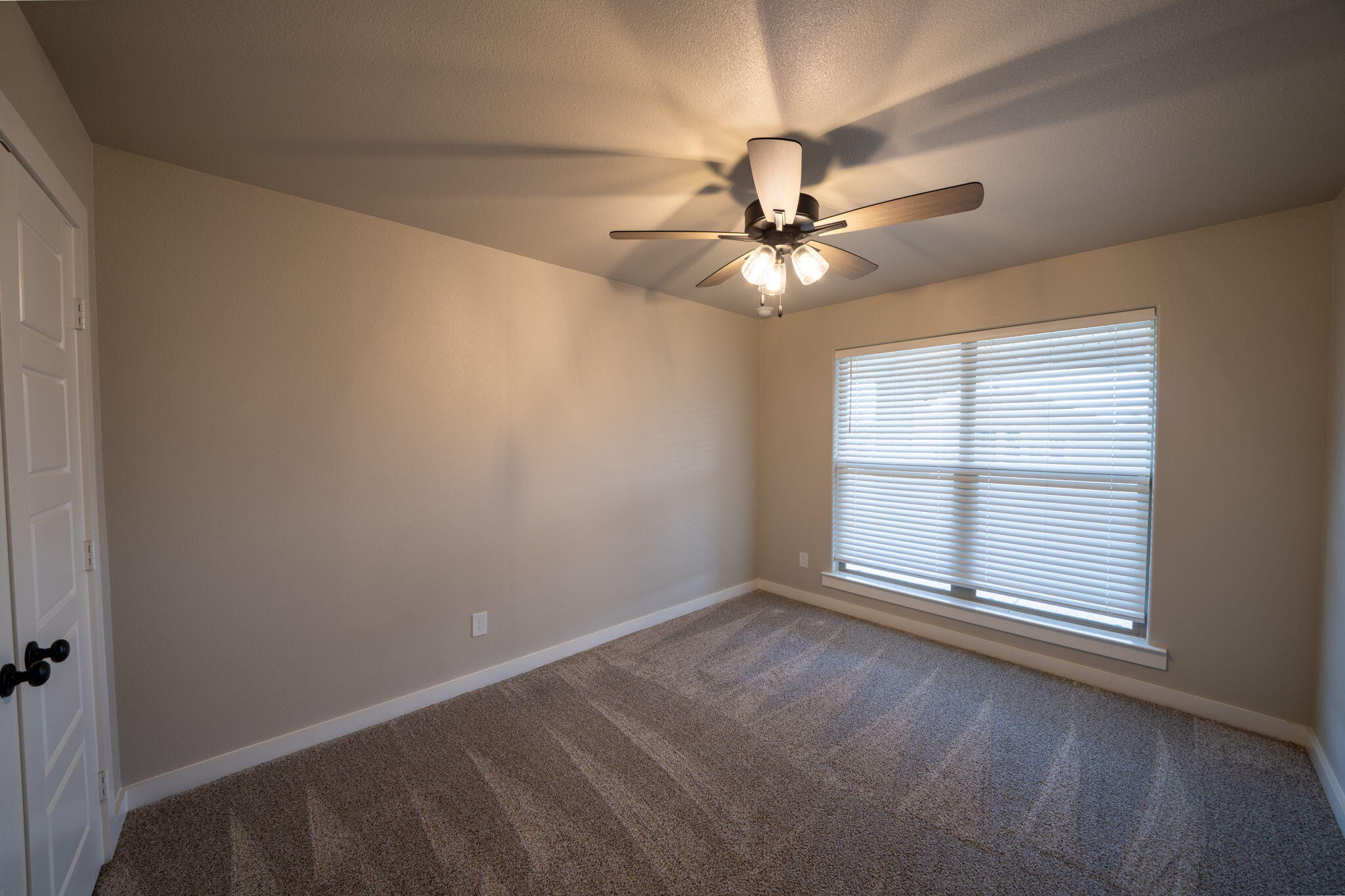 Empty carpeted bedroom with a ceiling fan, white closet doors, and a large window with blinds.