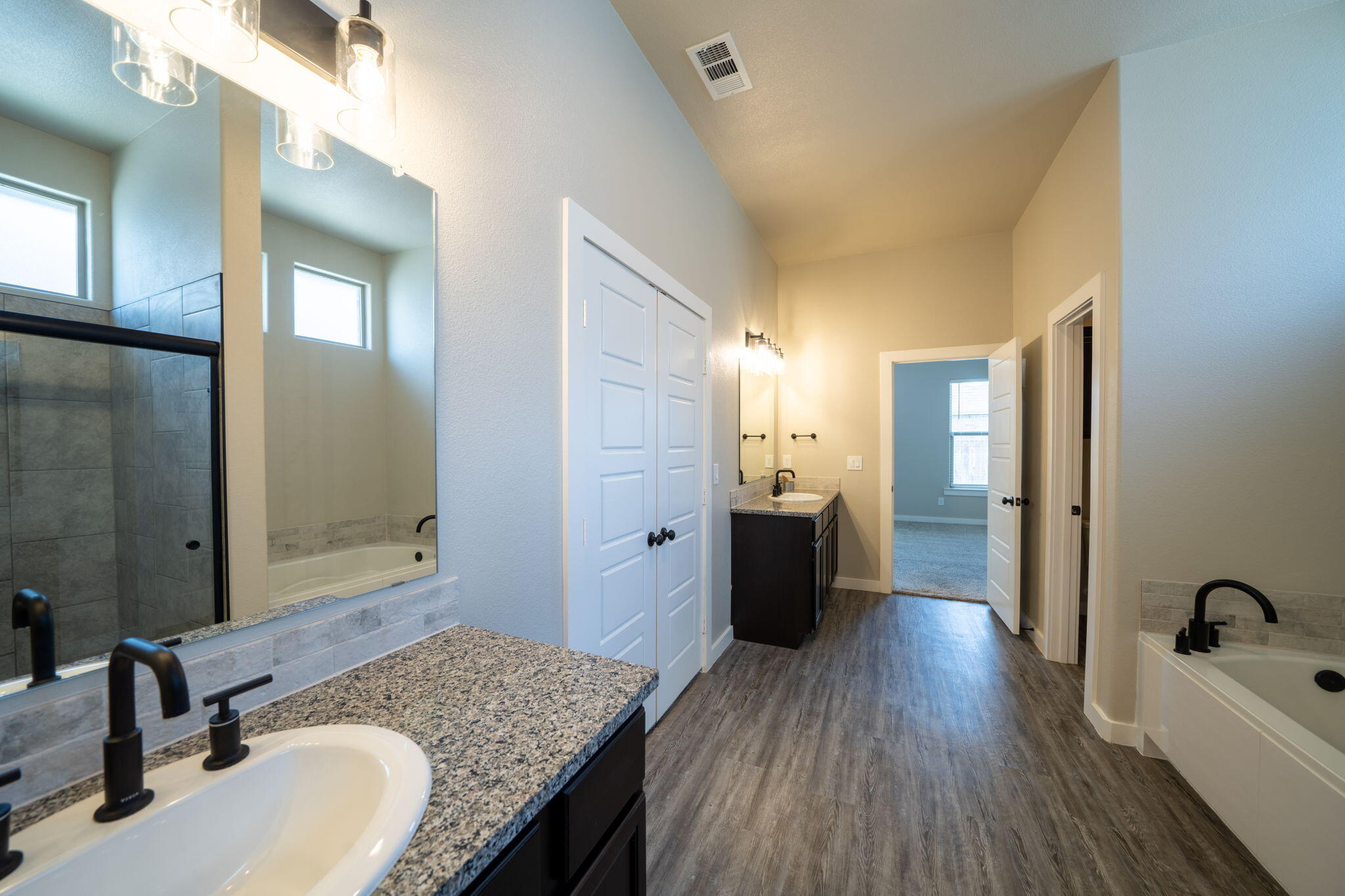 Long bathroom with double vanities, granite counters, tiled shower, and wood-look flooring.