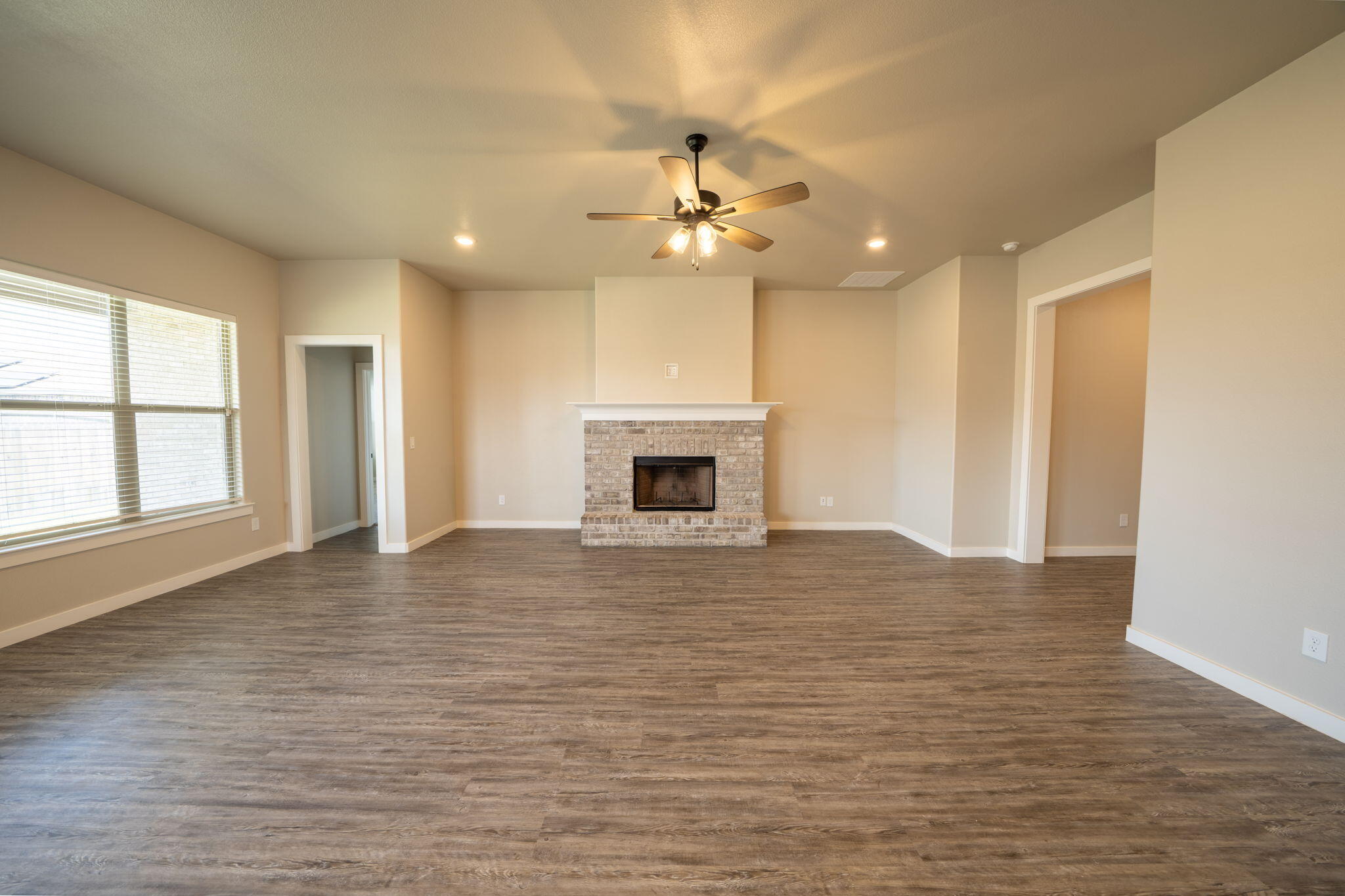 Empty living room with a brick fireplace, wood-look floors, ceiling fan, and large window.