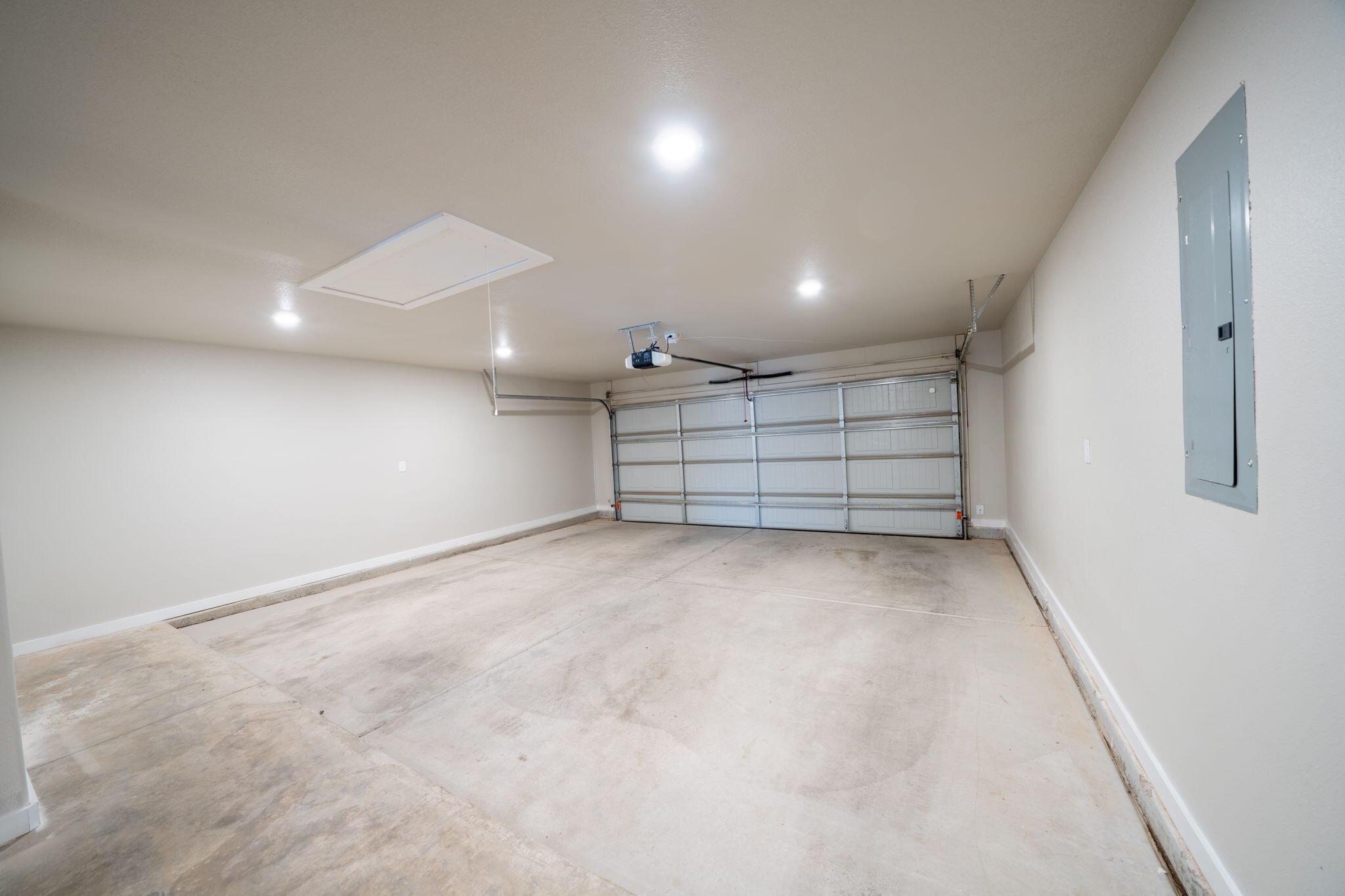 Empty garage with concrete floor, closed sectional door, ceiling-mounted opener, and wall electrical panel.