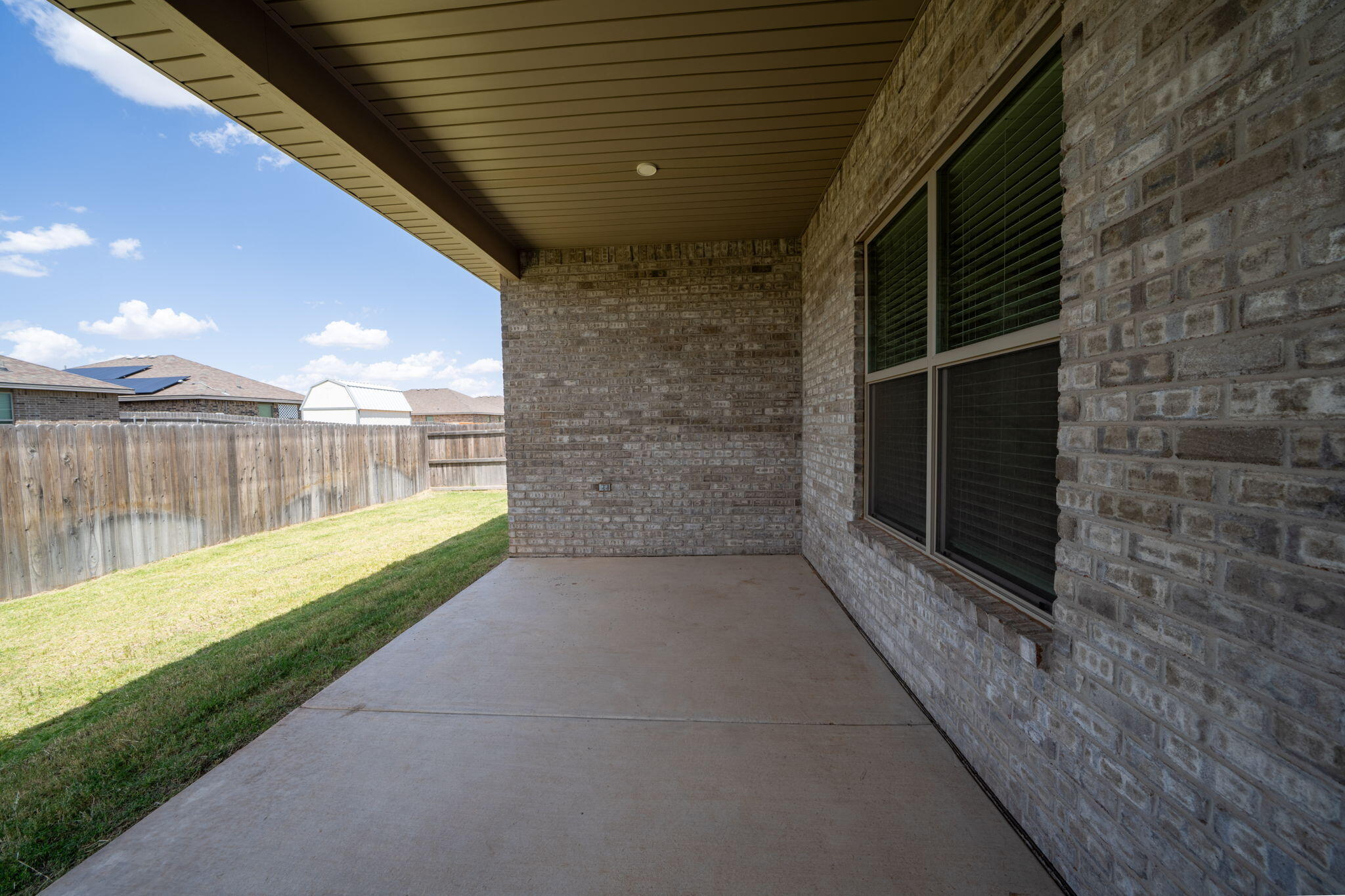 Covered concrete patio beside a brick house, overlooking a fenced grassy backyard.