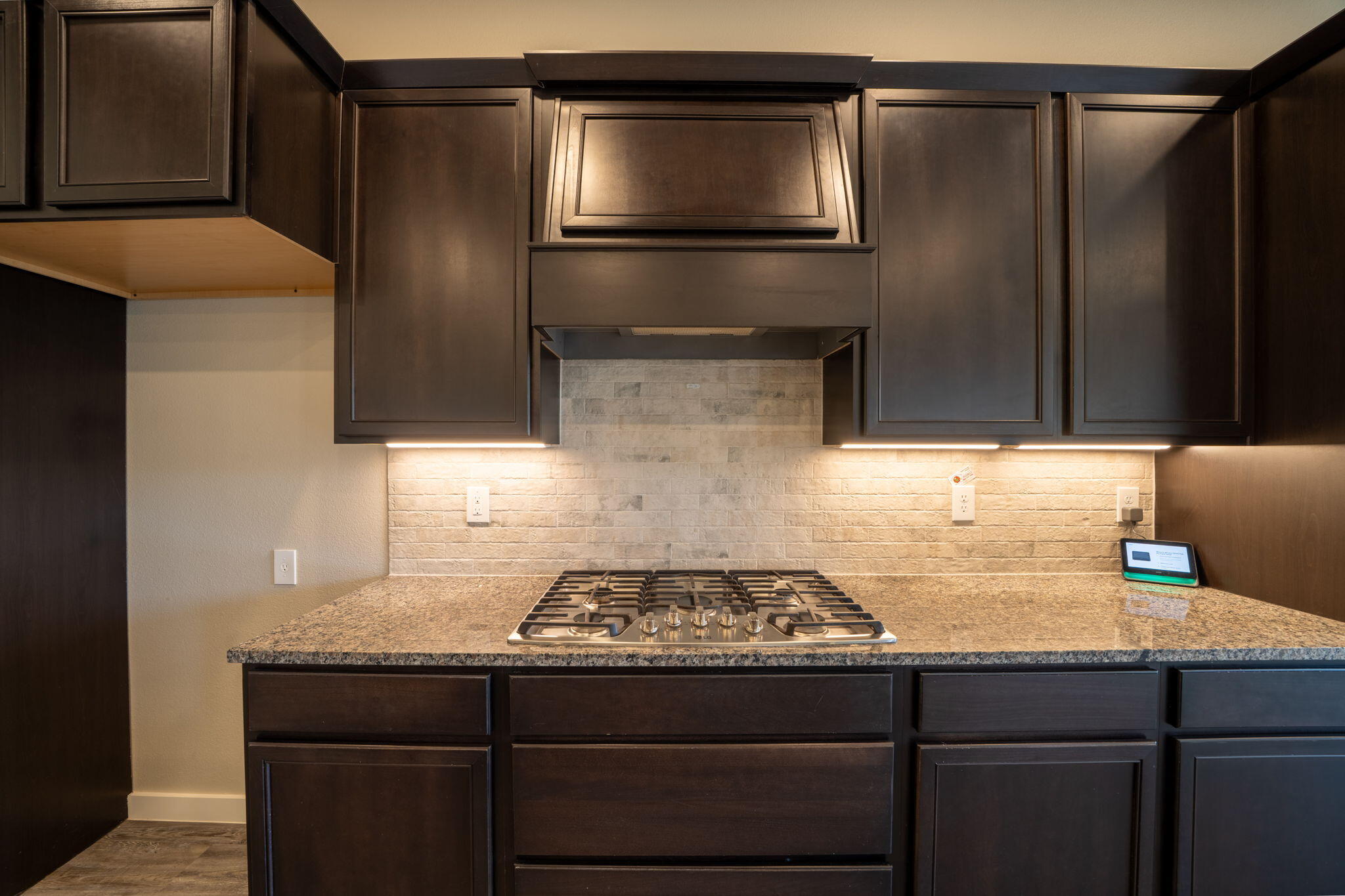 Kitchen cooktop beneath dark wood cabinets, with a light brick backsplash and granite counter.