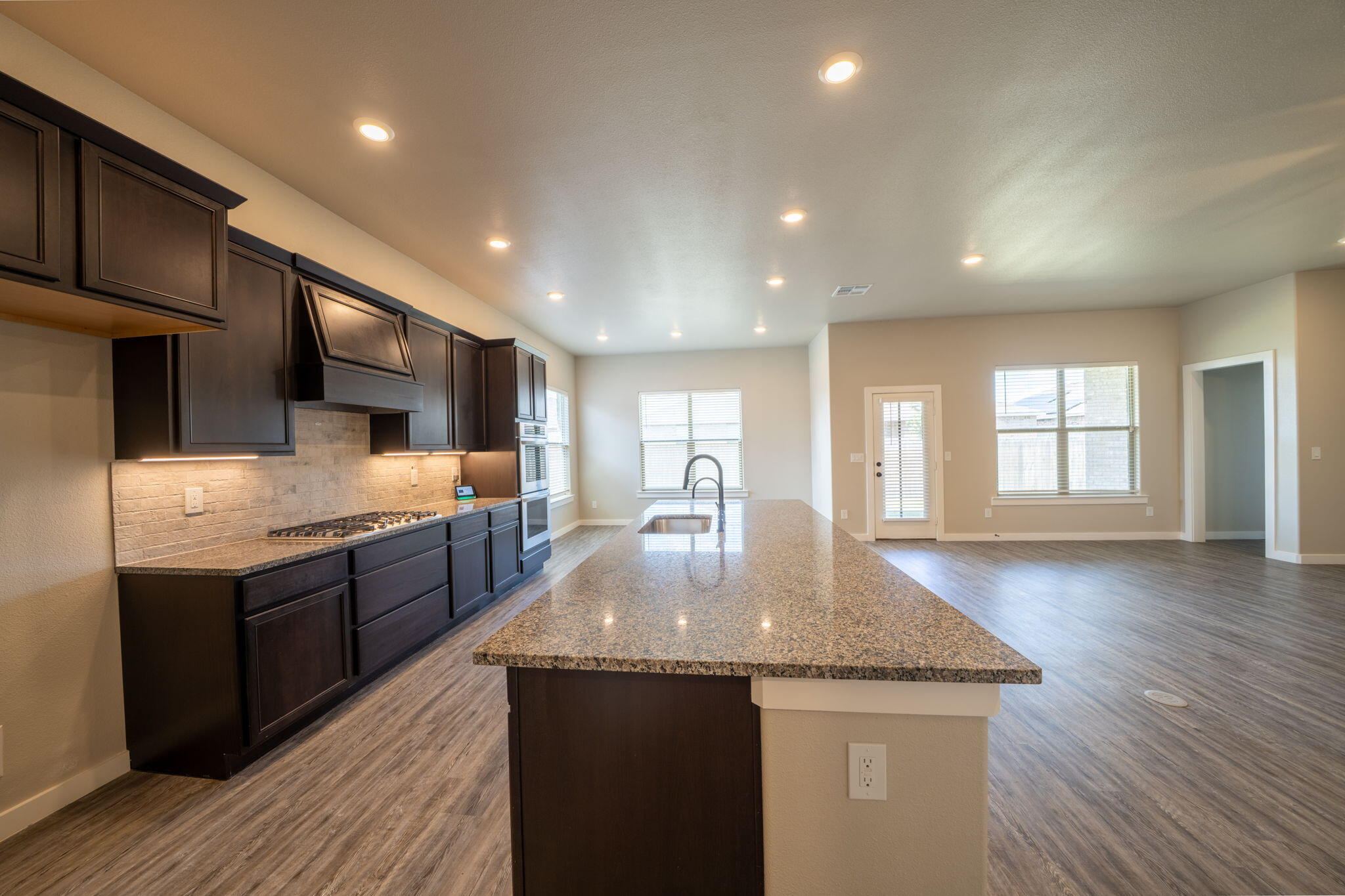 Open kitchen with dark cabinets, granite island, stainless steel appliances, and adjoining unfurnished living area.