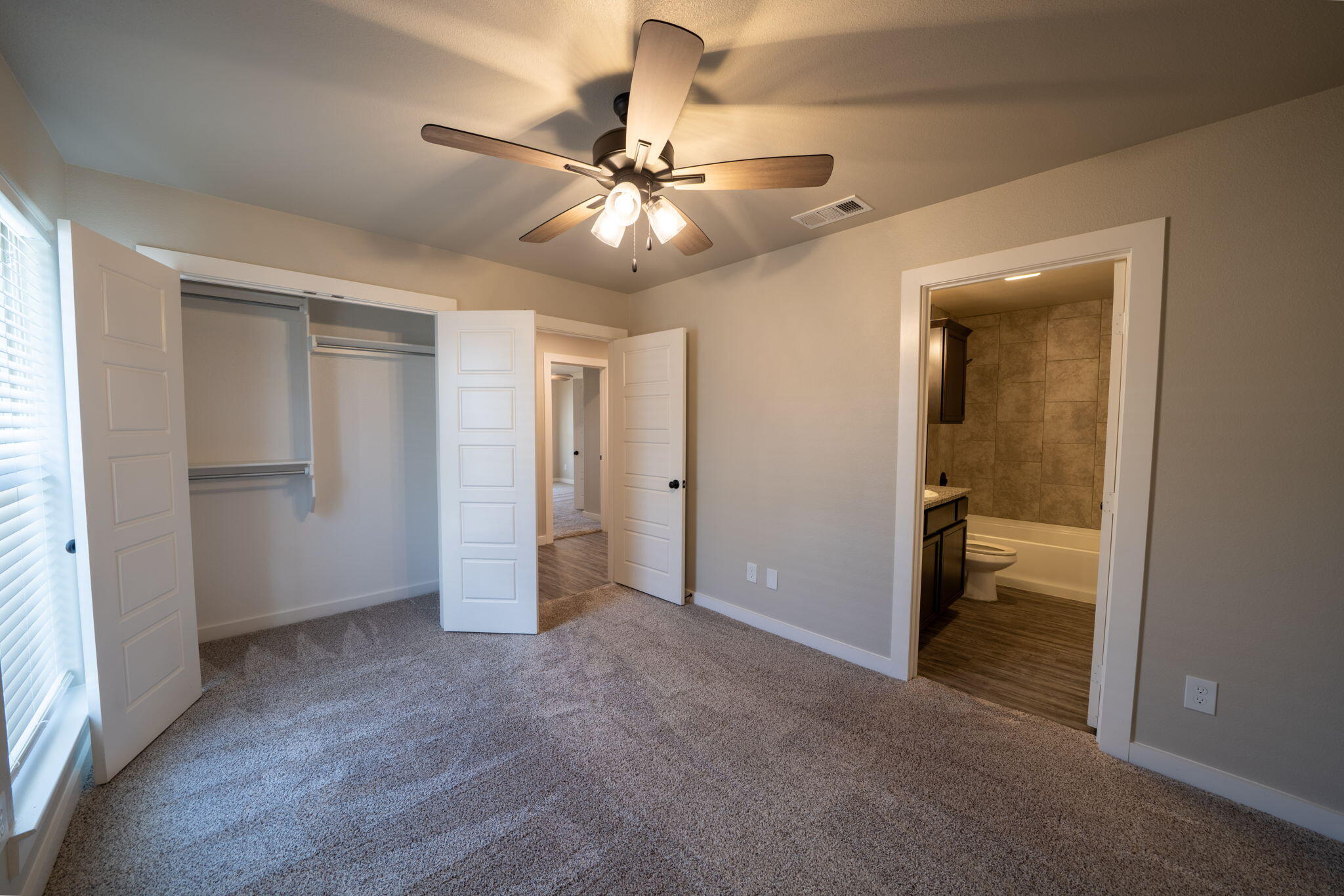 Empty carpeted bedroom with an open closet, ceiling fan, and doorway to an ensuite bathroom.