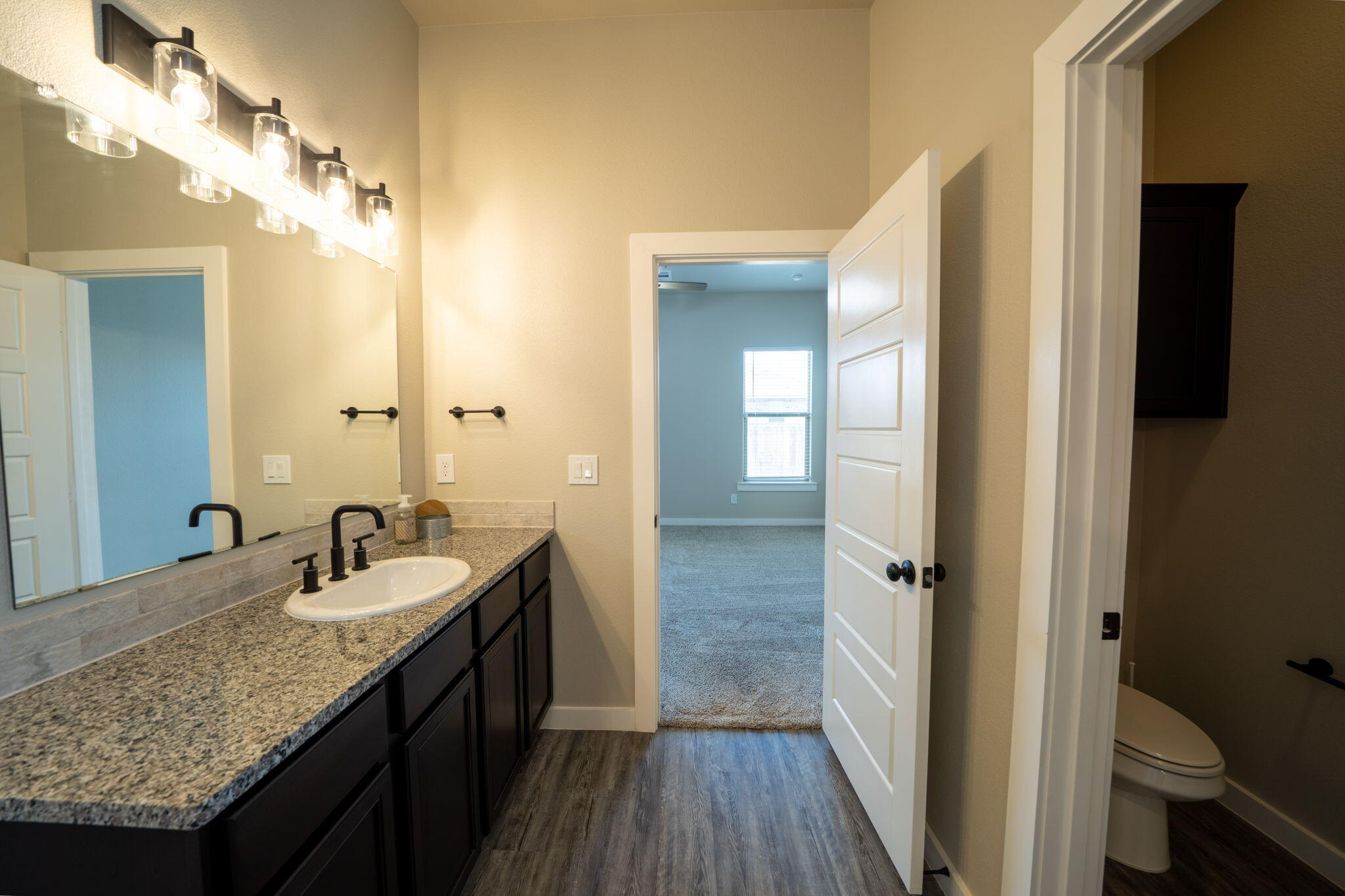 Bathroom with granite vanity, dark cabinets, bright mirror lights, and open doors to a toilet room and bedroom.