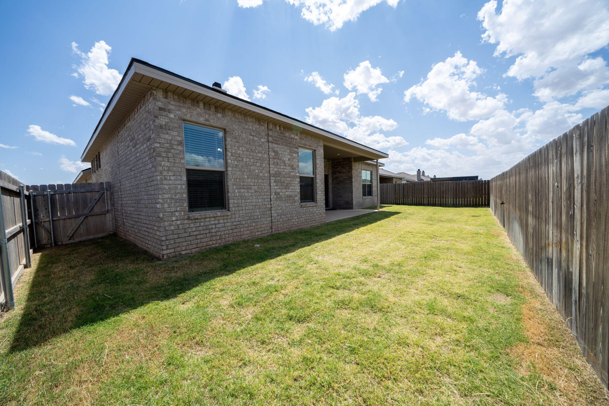 Brick house and covered patio beside a grassy backyard enclosed by wooden privacy fencing.