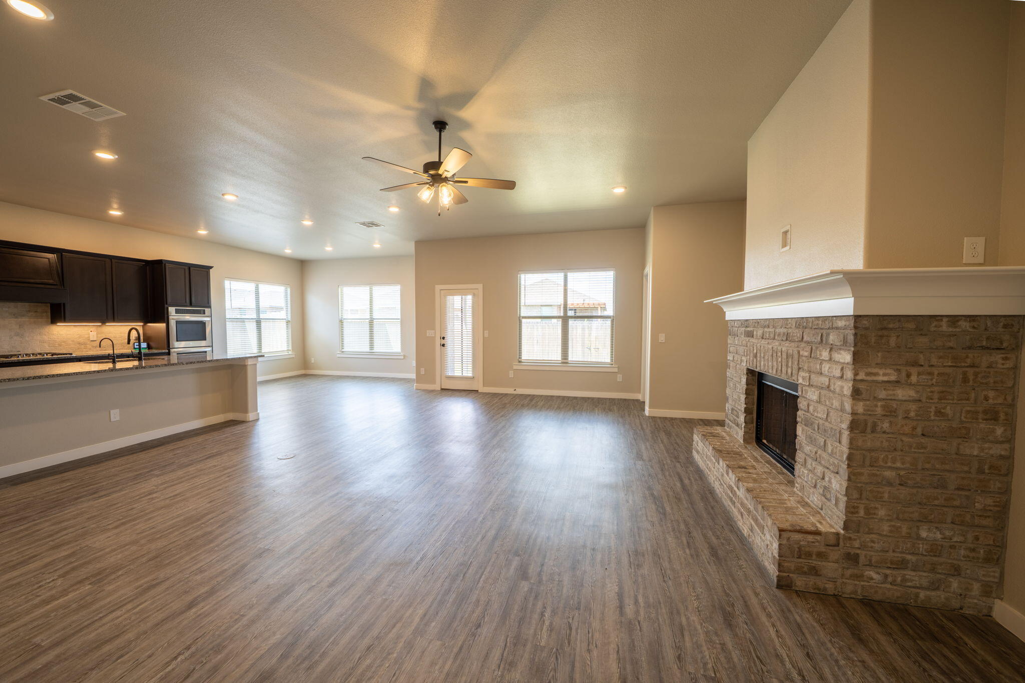 Empty open-plan living room with dark kitchen cabinets, wood-look floors, and a large brick fireplace.