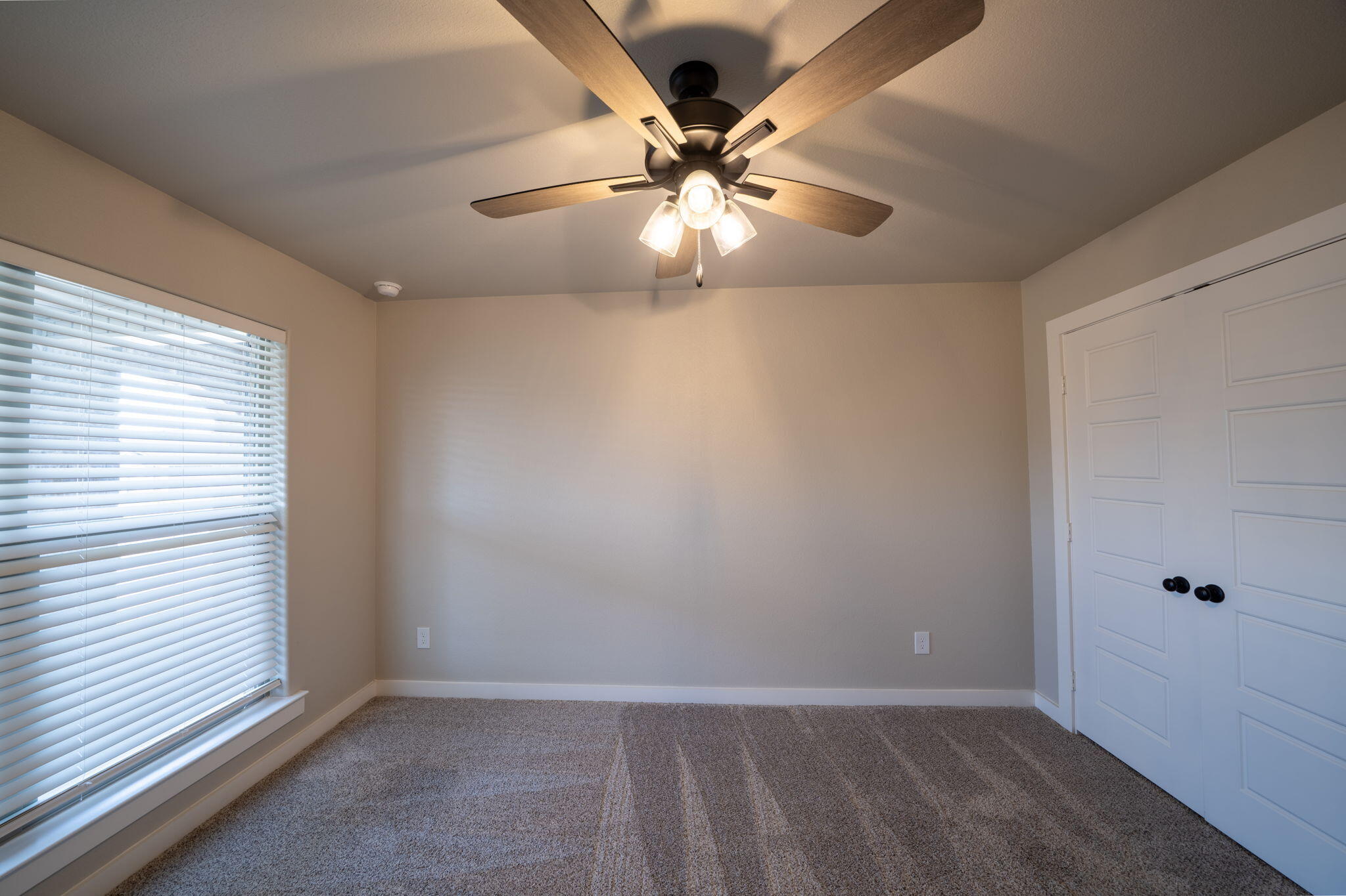 Empty bedroom with gray carpet, window blinds, double closet doors, and a lit ceiling fan.