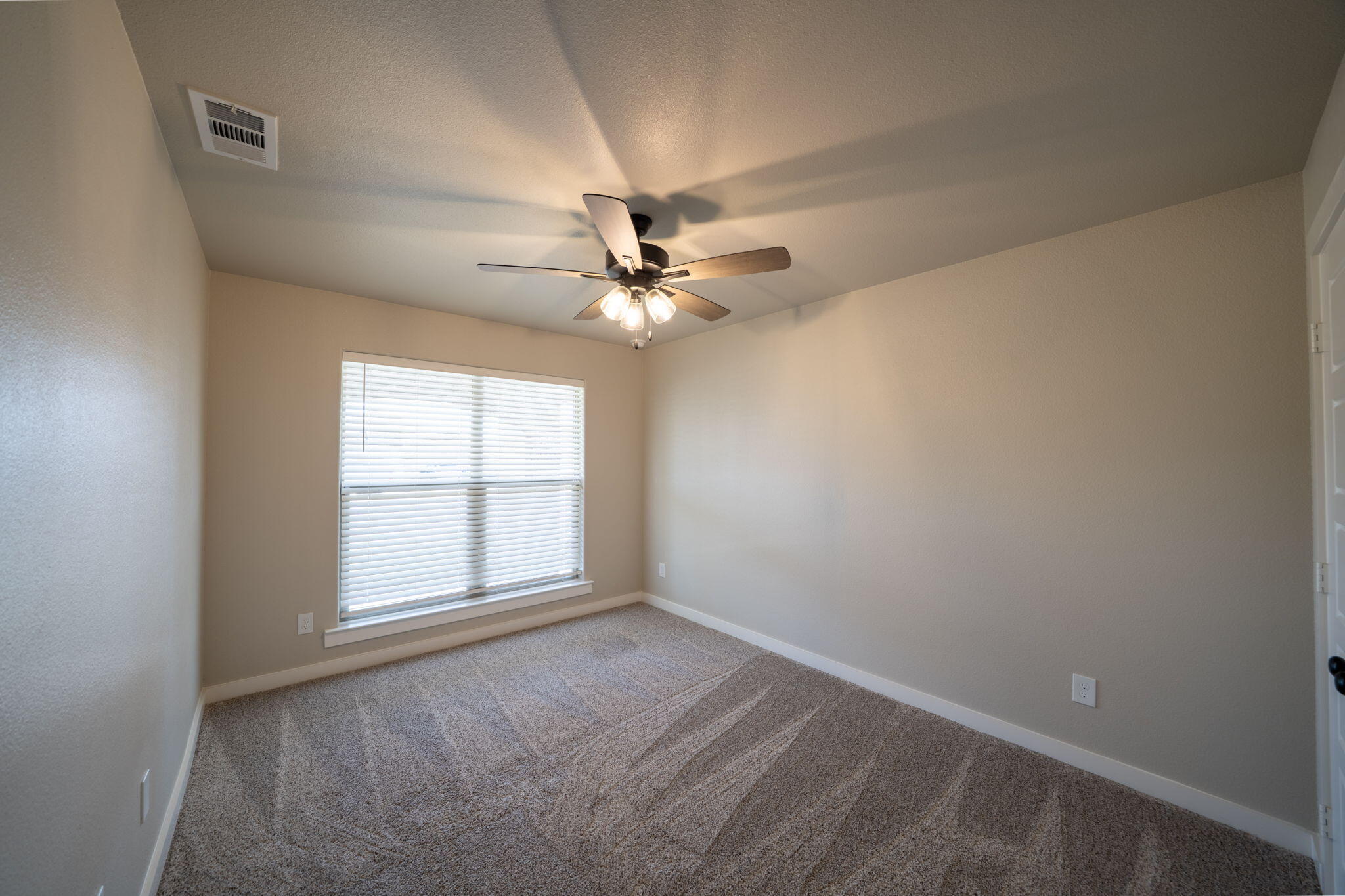 Empty bedroom with gray carpet, a wide window with blinds, and a lit ceiling fan.
