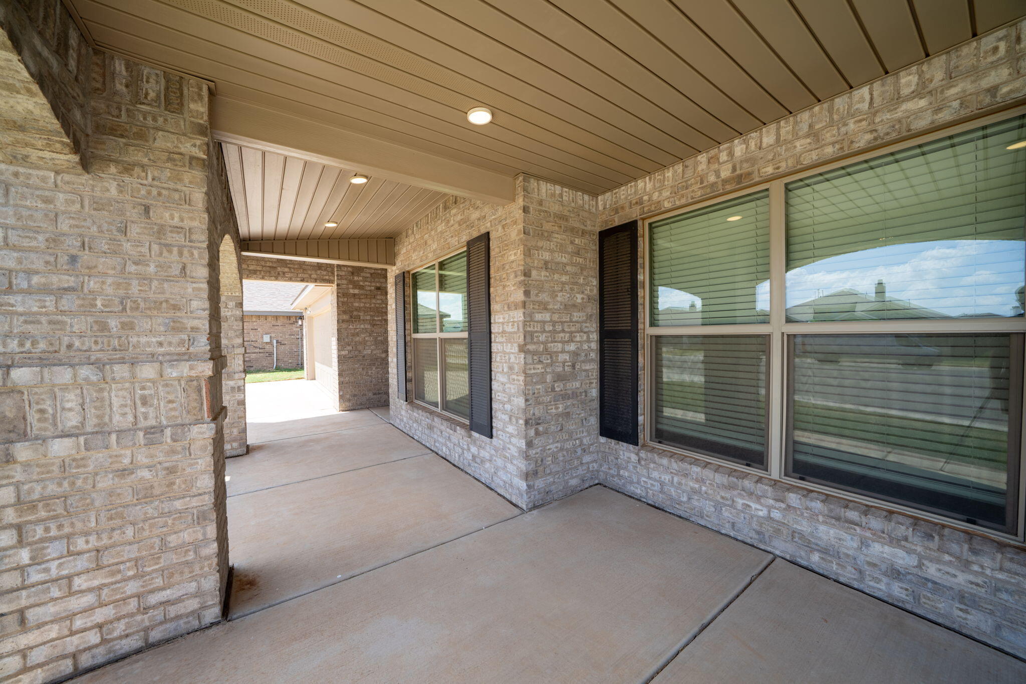 Covered concrete patio beside a light brick house, with large windows and an open passage to the side yard.