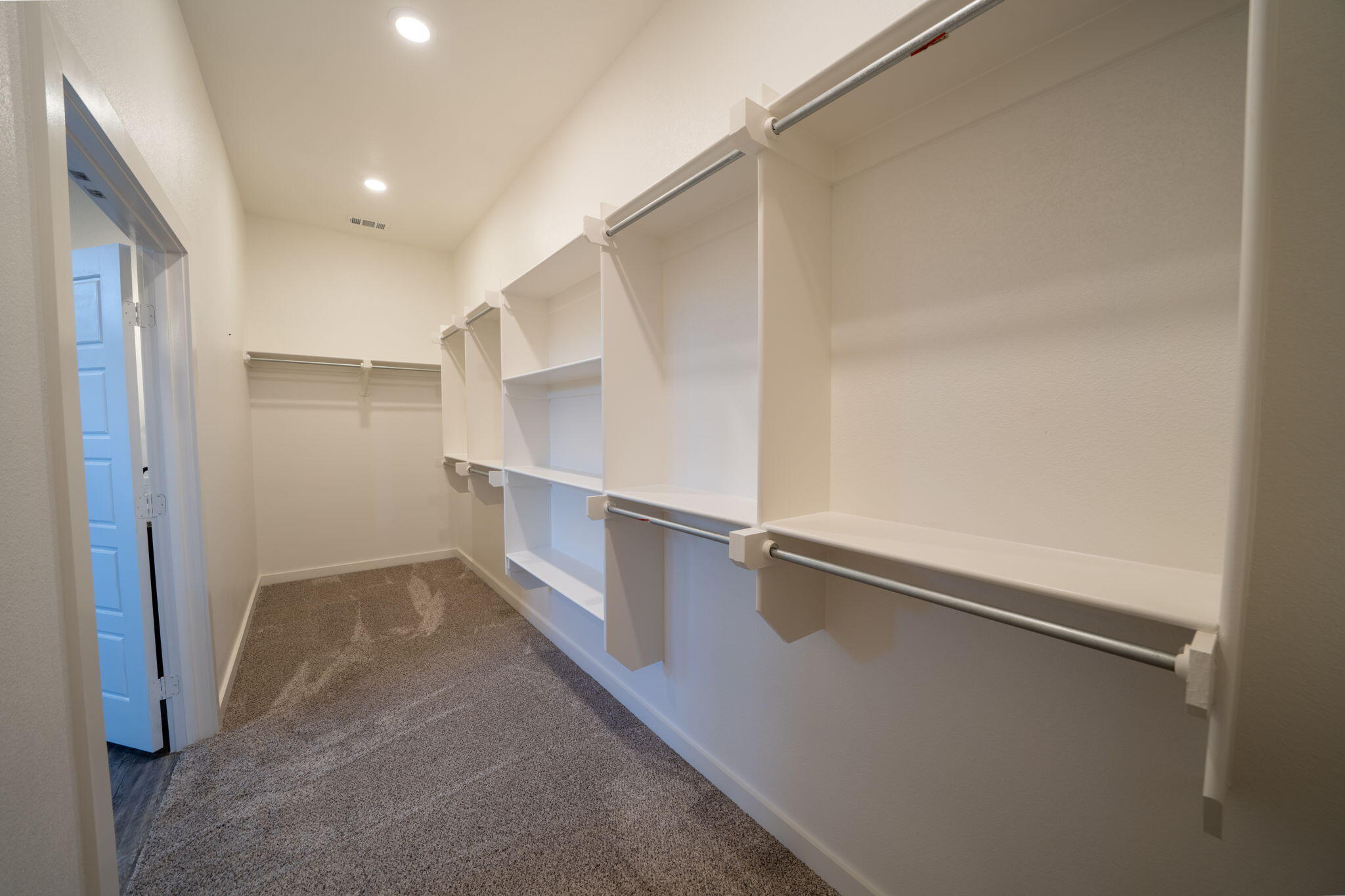 Walk-in closet with white shelving and clothes rods above beige carpet.