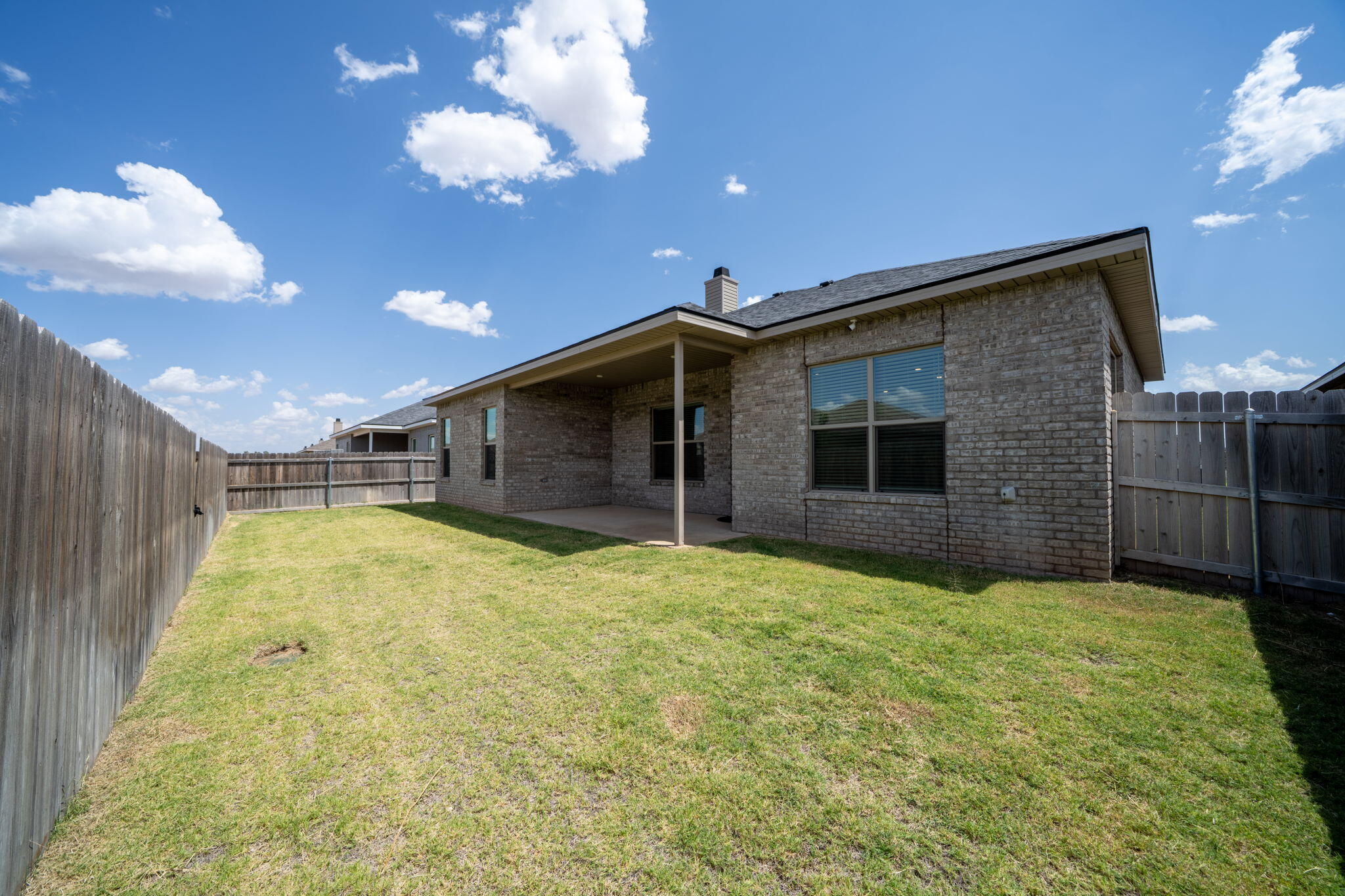 Brick house and covered patio behind a fenced grassy backyard.