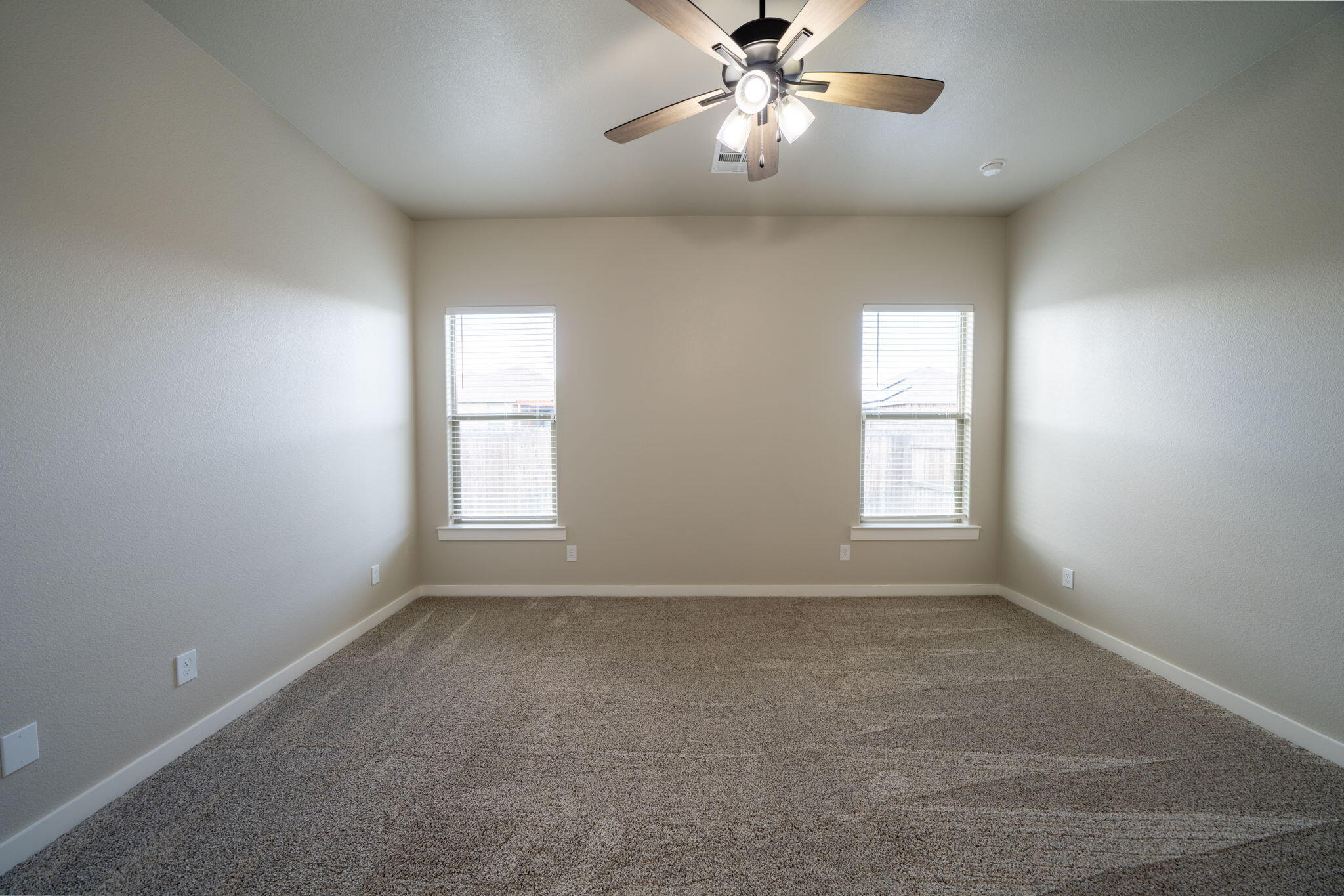 Empty carpeted bedroom with two blinded windows and a ceiling fan with lights.