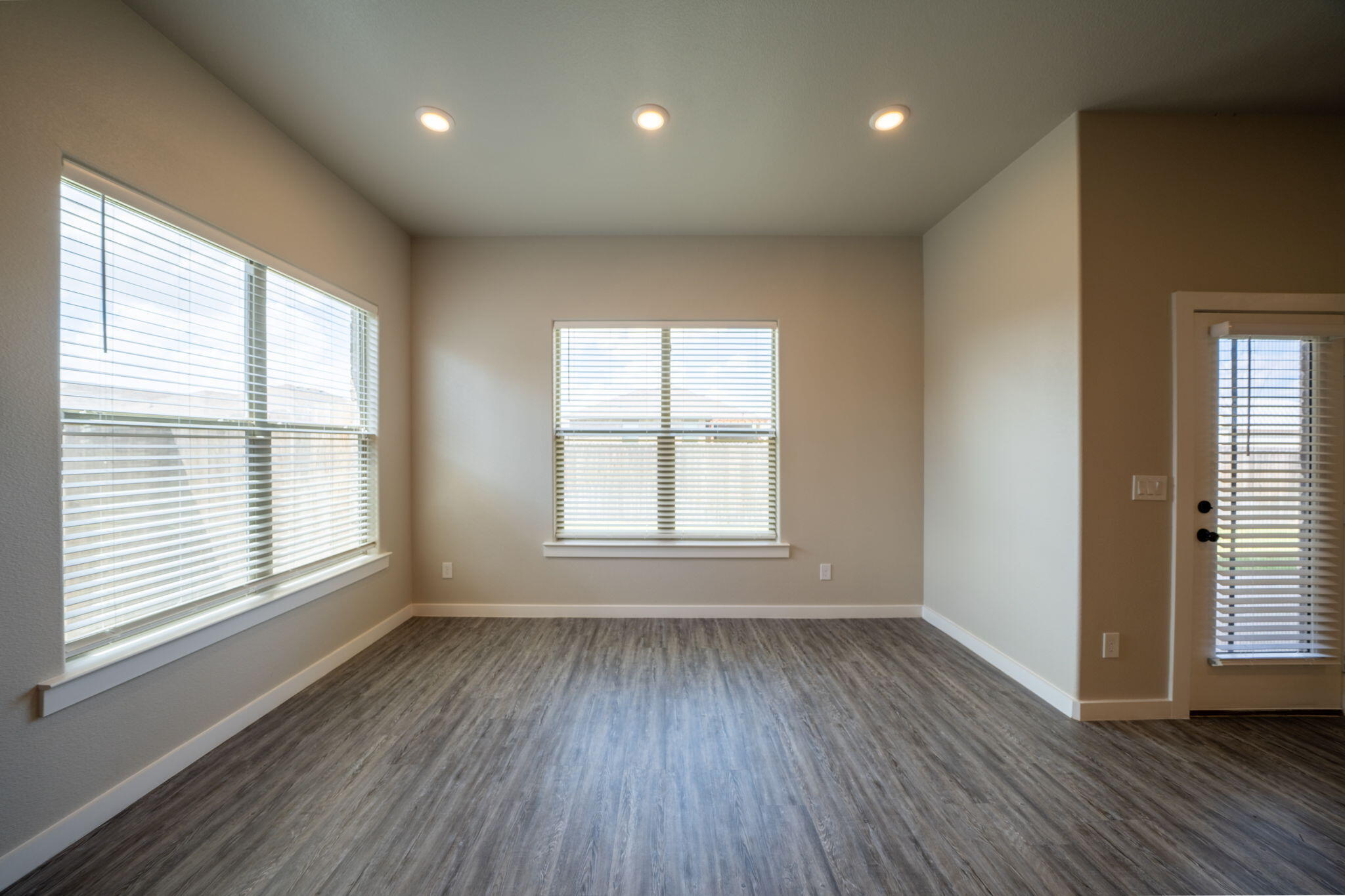 Empty living area with gray wood-look flooring, recessed lights, and windows with blinds.