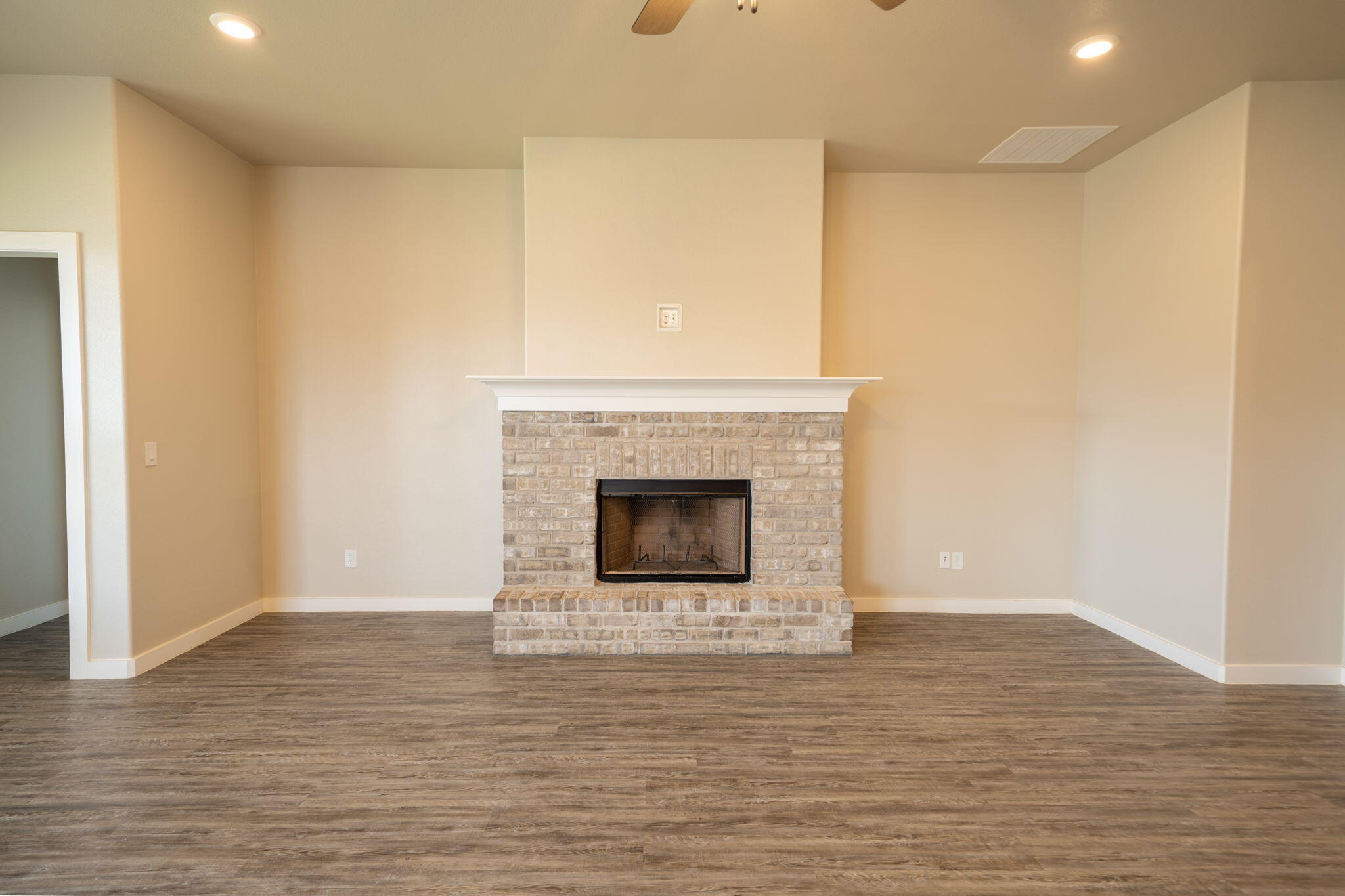 Empty living room with a light brick fireplace, white mantel, and brown wood-look flooring.