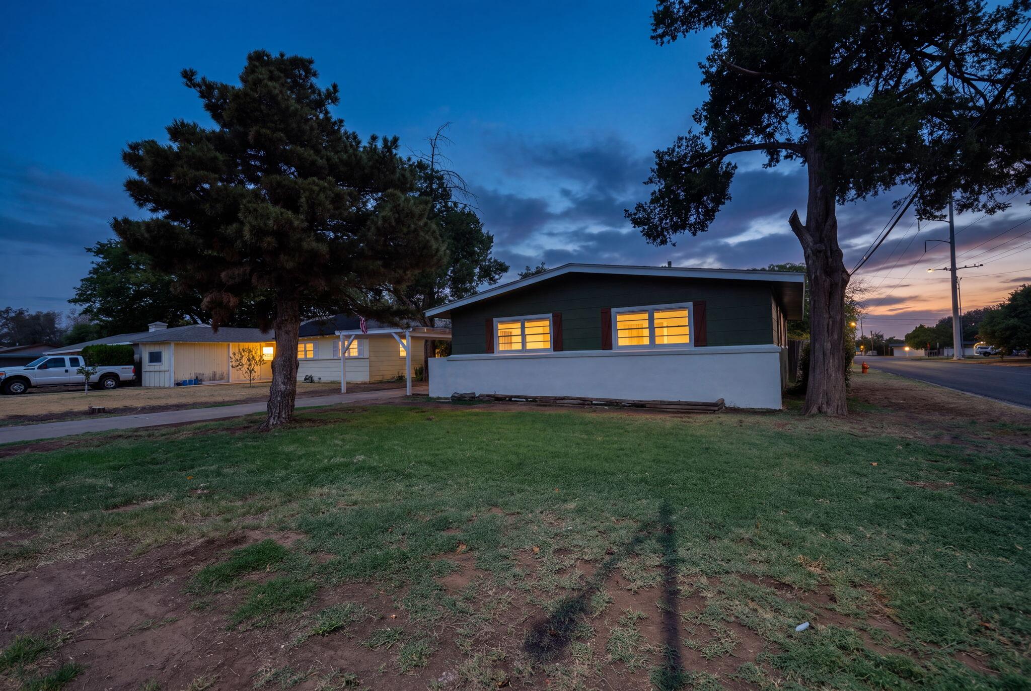 Single-story dark green house with warmly lit windows, framed by large trees at dusk.