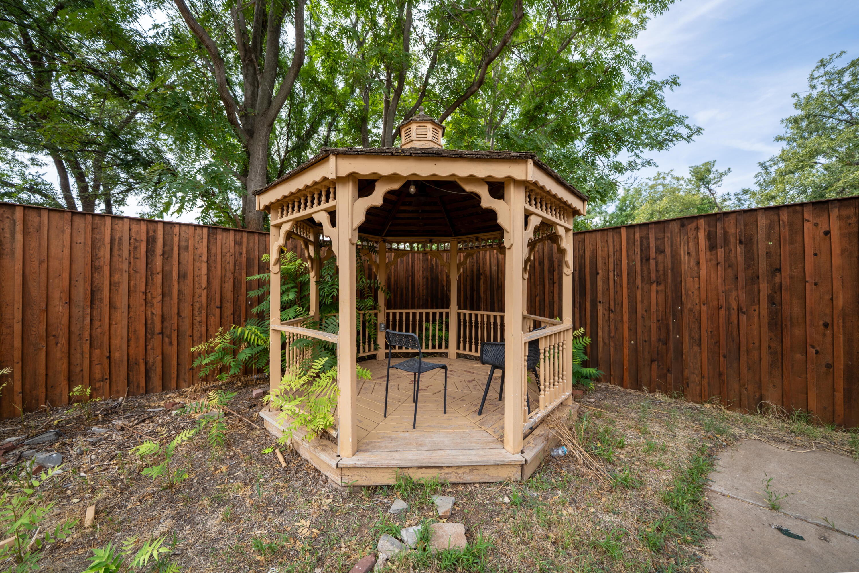Wooden backyard gazebo with decorative trim, two chairs, and mature trees.