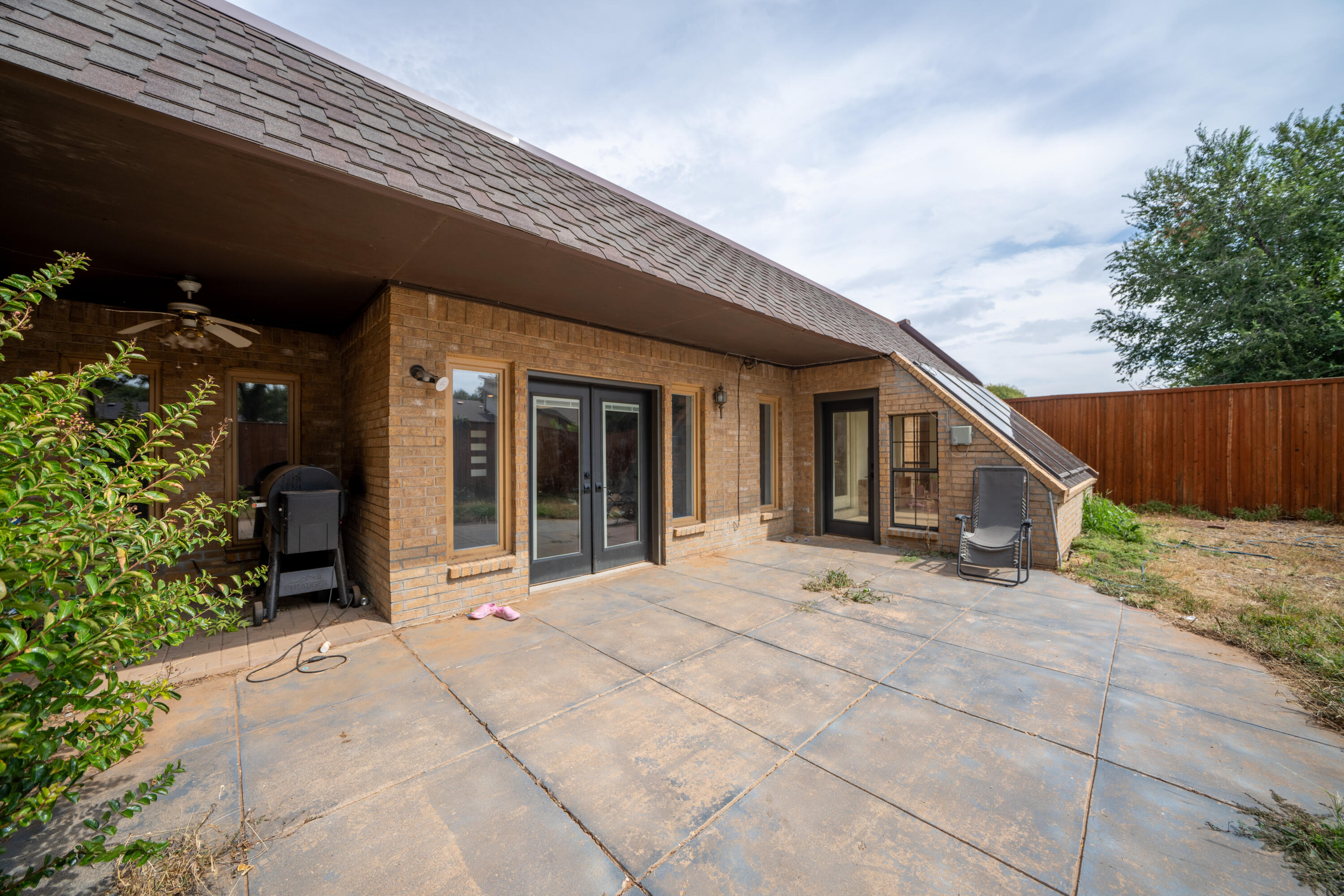 Brick home’s covered patio with glass doors, a concrete-paved seating area, and fenced yard.