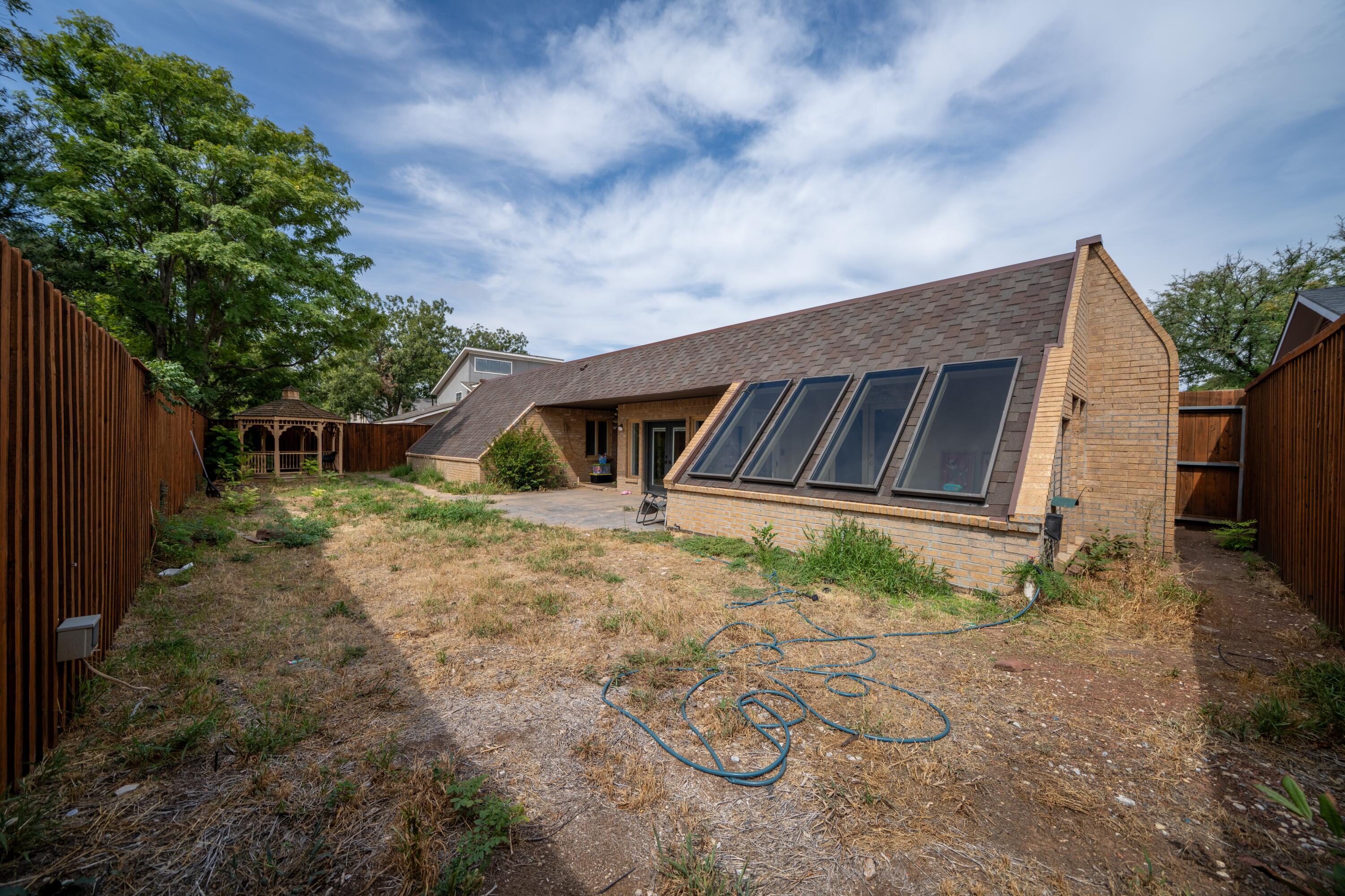 Fenced backyard with a brick home, large slanted roof windows, dry grass, and a wooden gazebo.