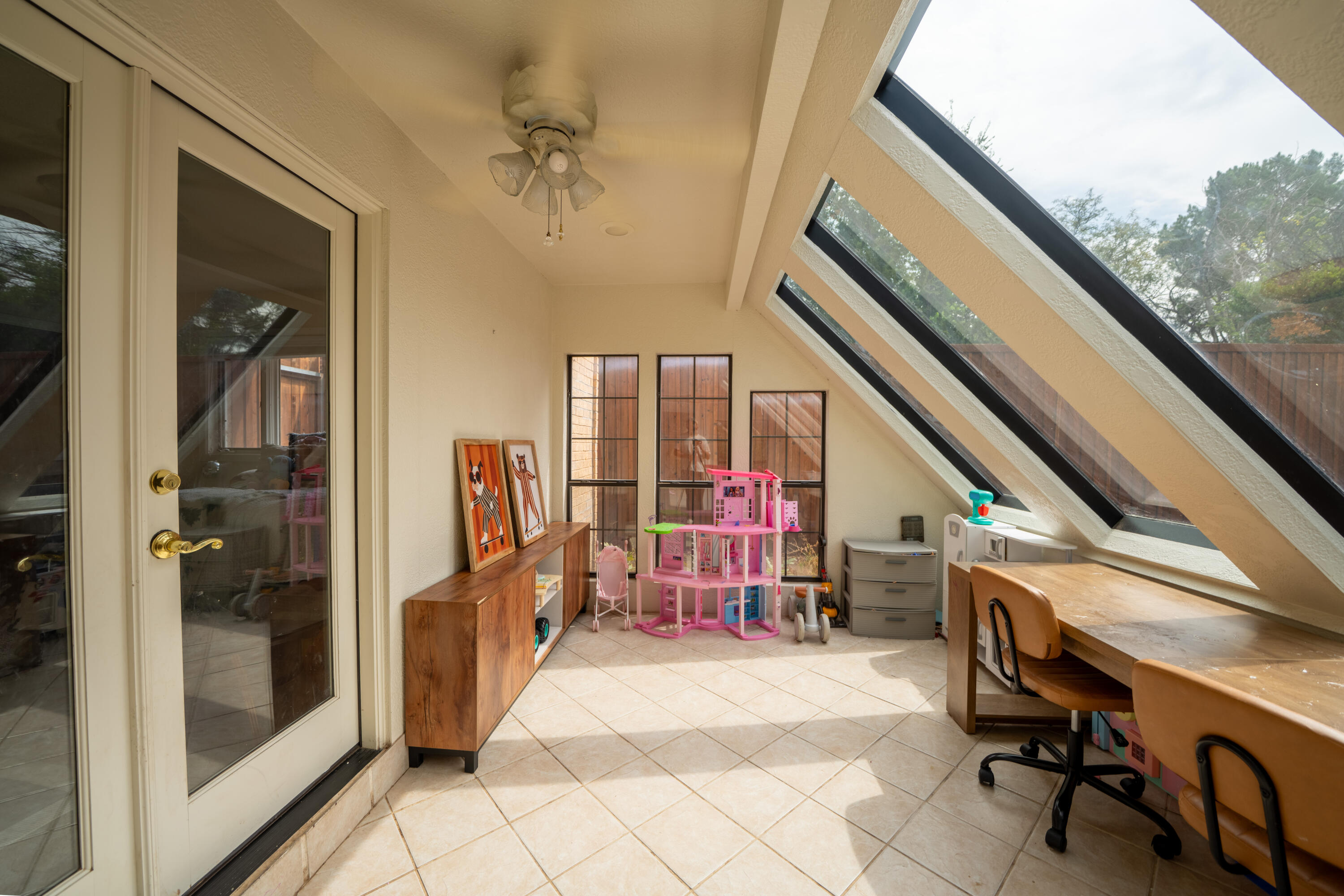 Sunroom with sloped windows, tiled floor, wooden desk, and children's toys.