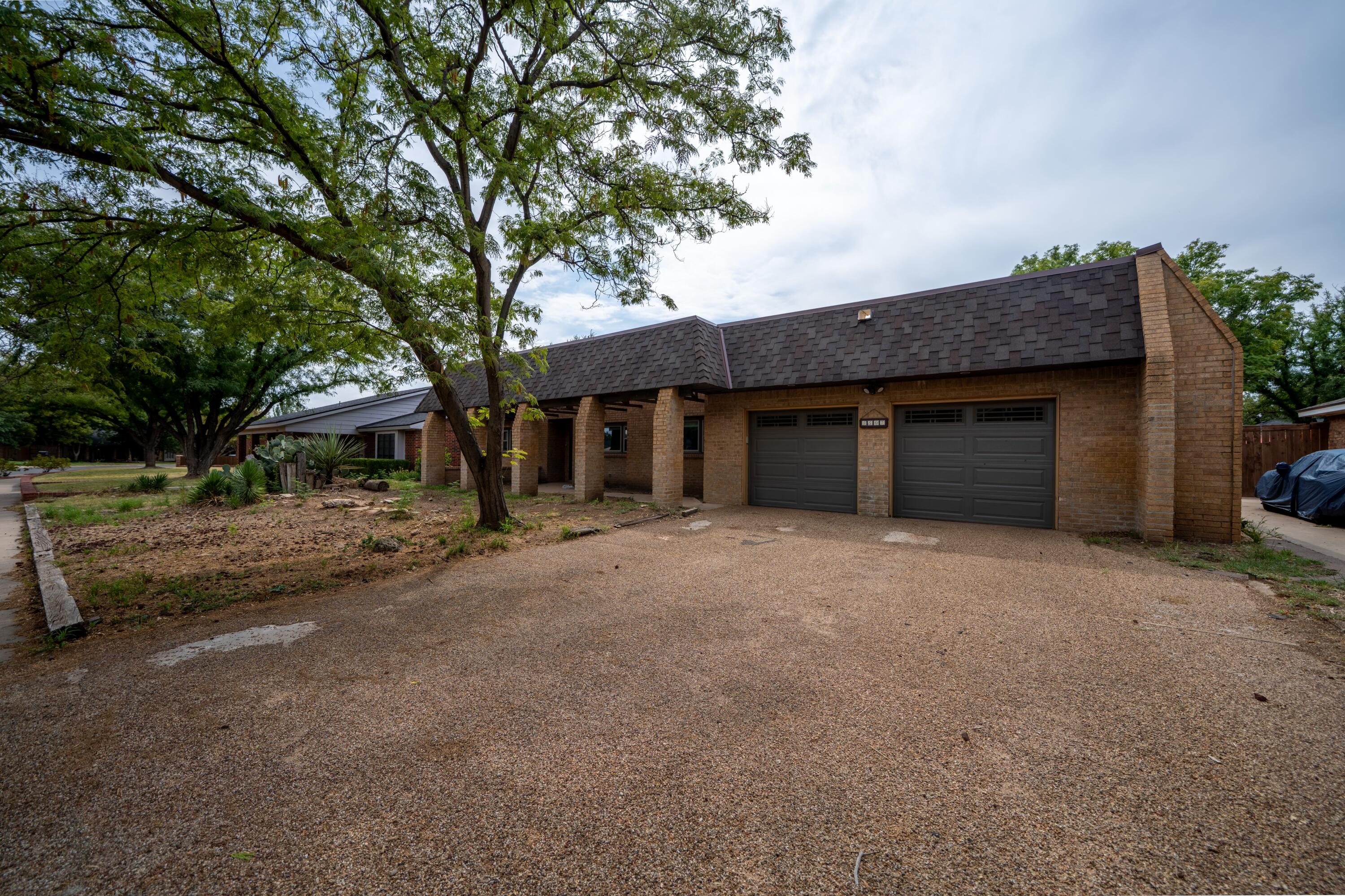 Brick home with a two-car garage, covered entrance, and gravel driveway beneath mature trees.