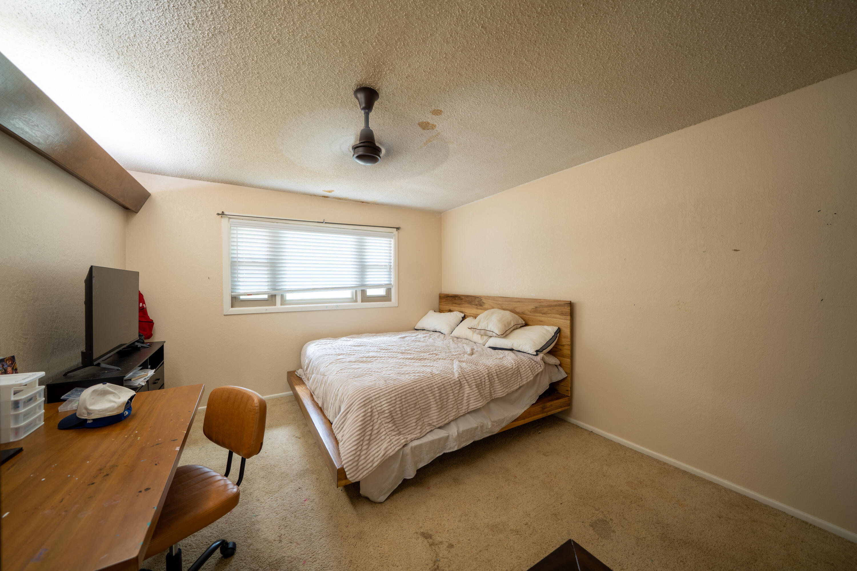 Bedroom with a wooden bed, window with blinds, and desk; ceiling has visible stains.