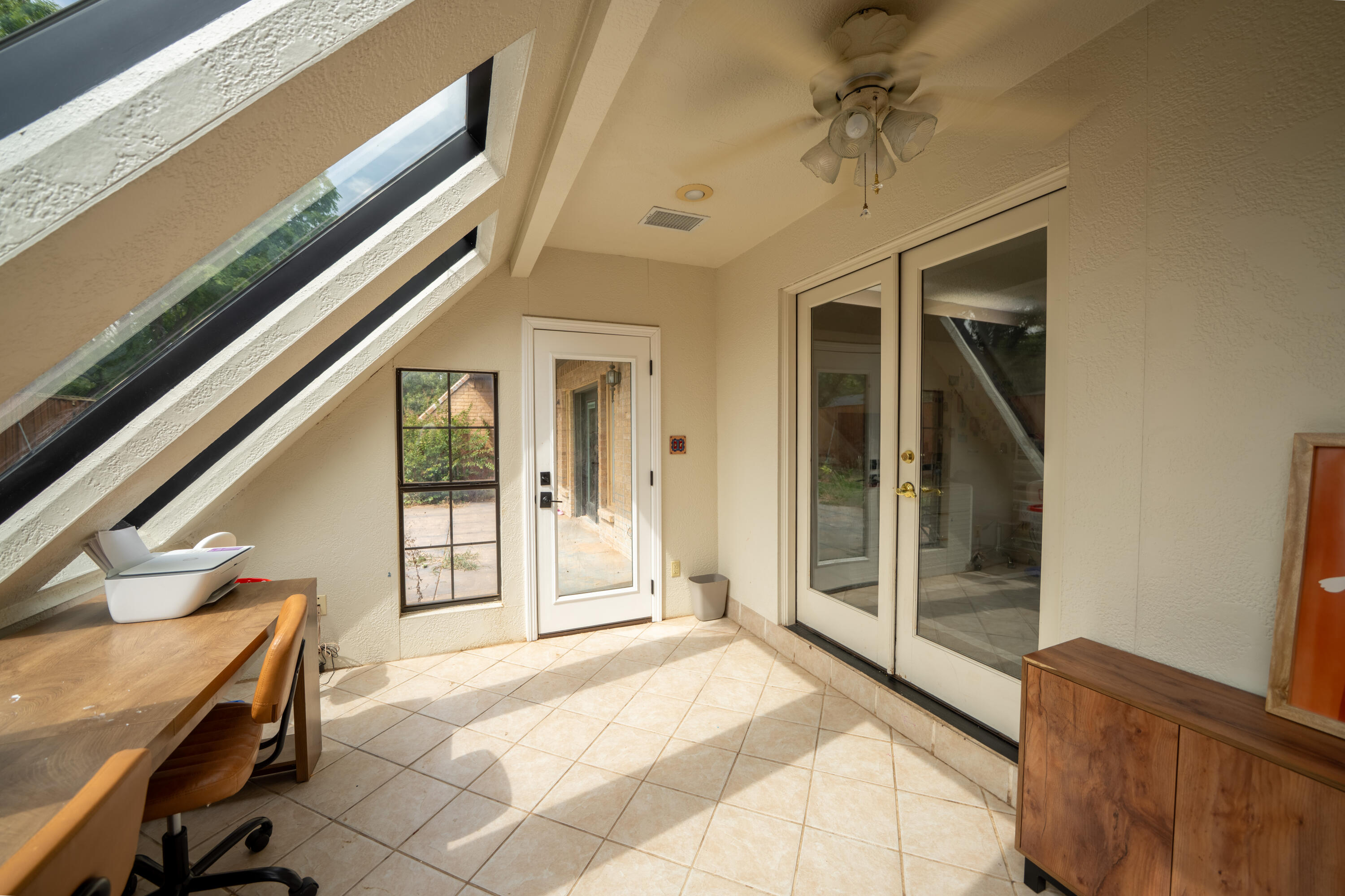 Sunroom with sloped windows, tiled floor, desk, and glass patio doors.