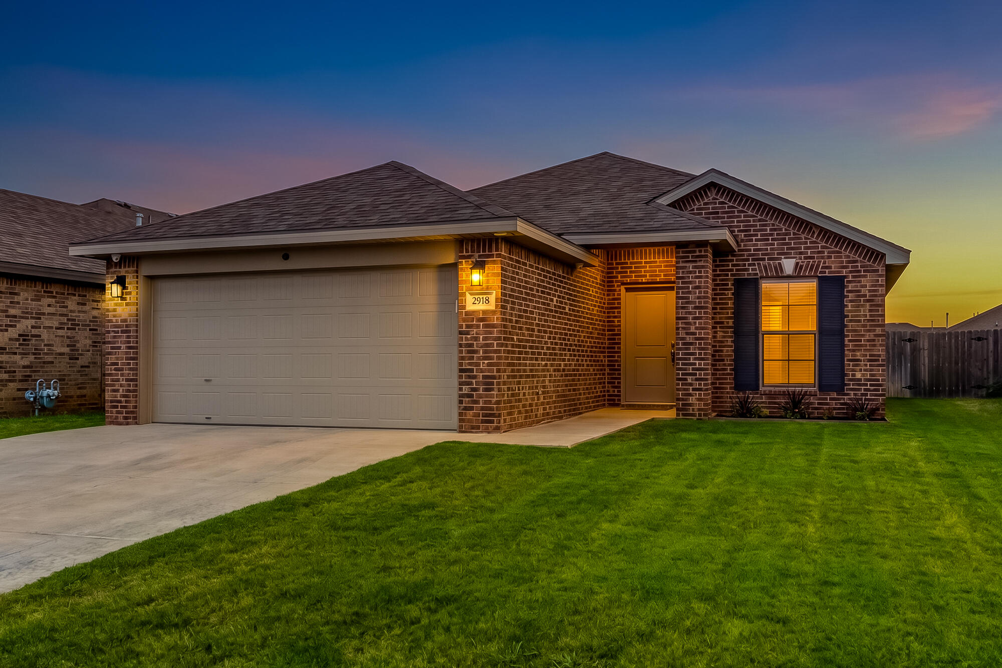 Brick house at dusk with a two-car garage, illuminated windows, and a broad green lawn.