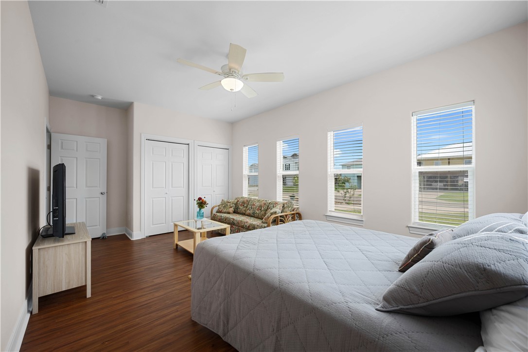 Furnished bedroom with a large bed, dark wood floors, and tall windows overlooking neighboring homes.