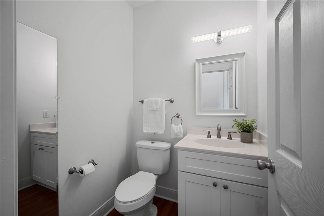 Bathroom with a white vanity and toilet, framed mirror, folded towels, and dark wood flooring beyond the doorway.