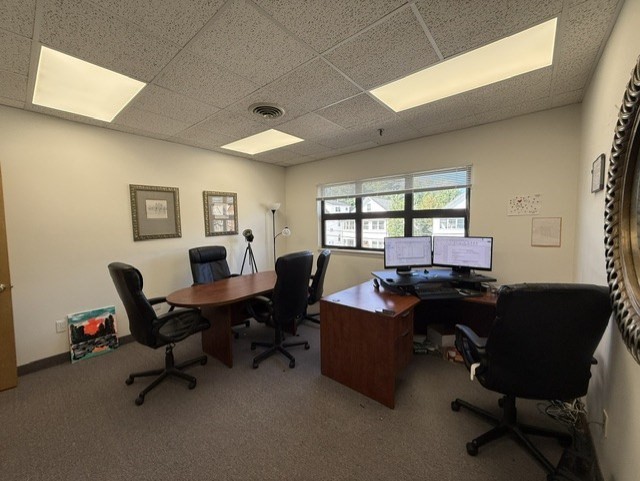 Furnished office with two desks, computer monitors, a conference table, and a large window.