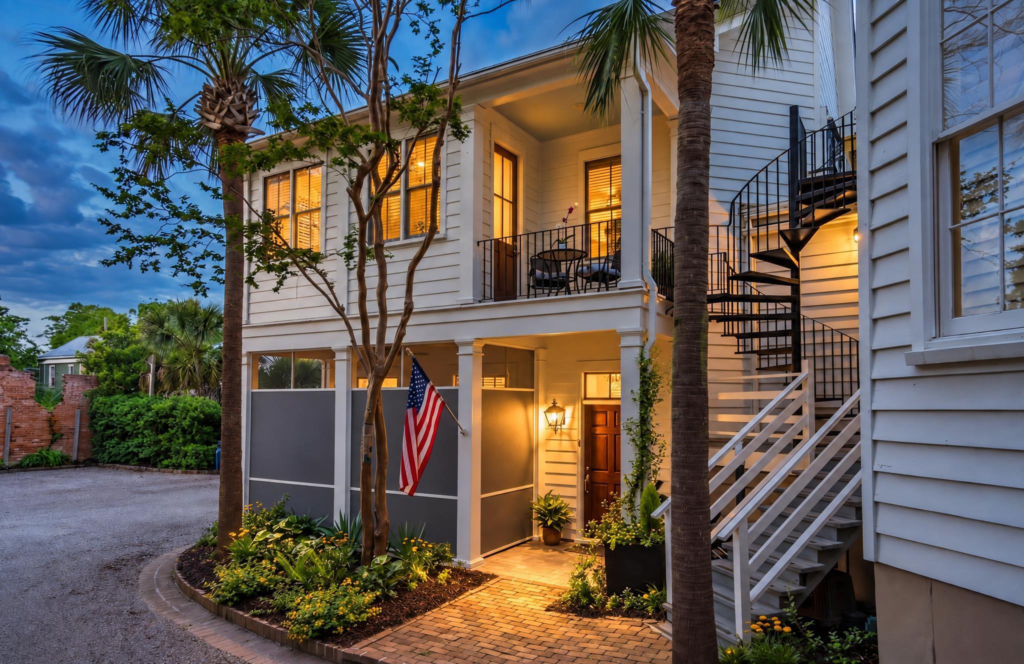 White-sided home with a warmly lit entry, spiral staircase, palm trees, and an American flag at dusk.
