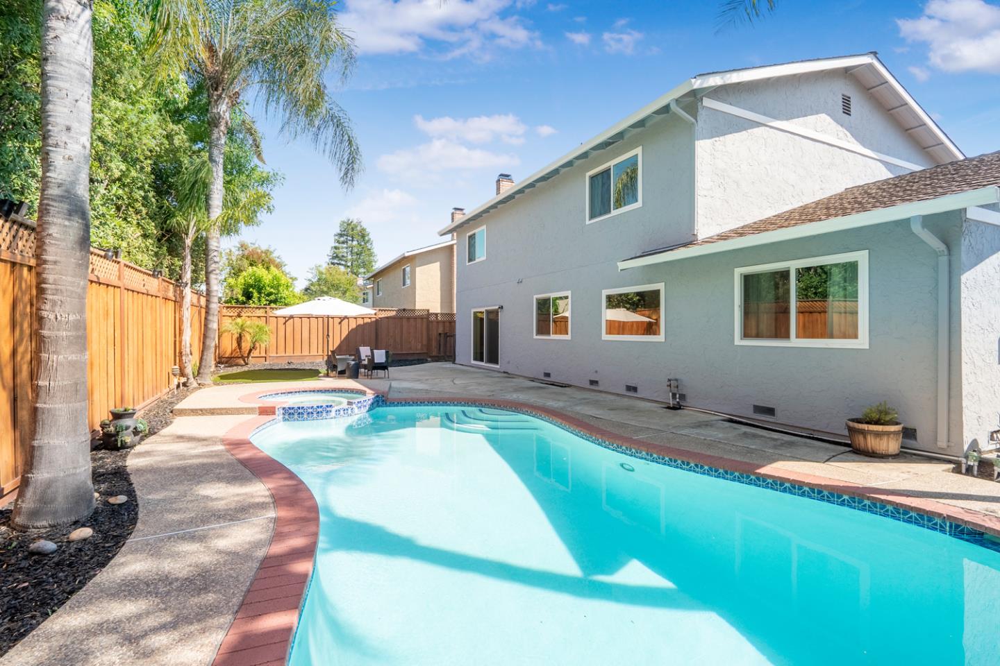 Backyard swimming pool beside a two-story stucco house, with palm trees and a wooden privacy fence.