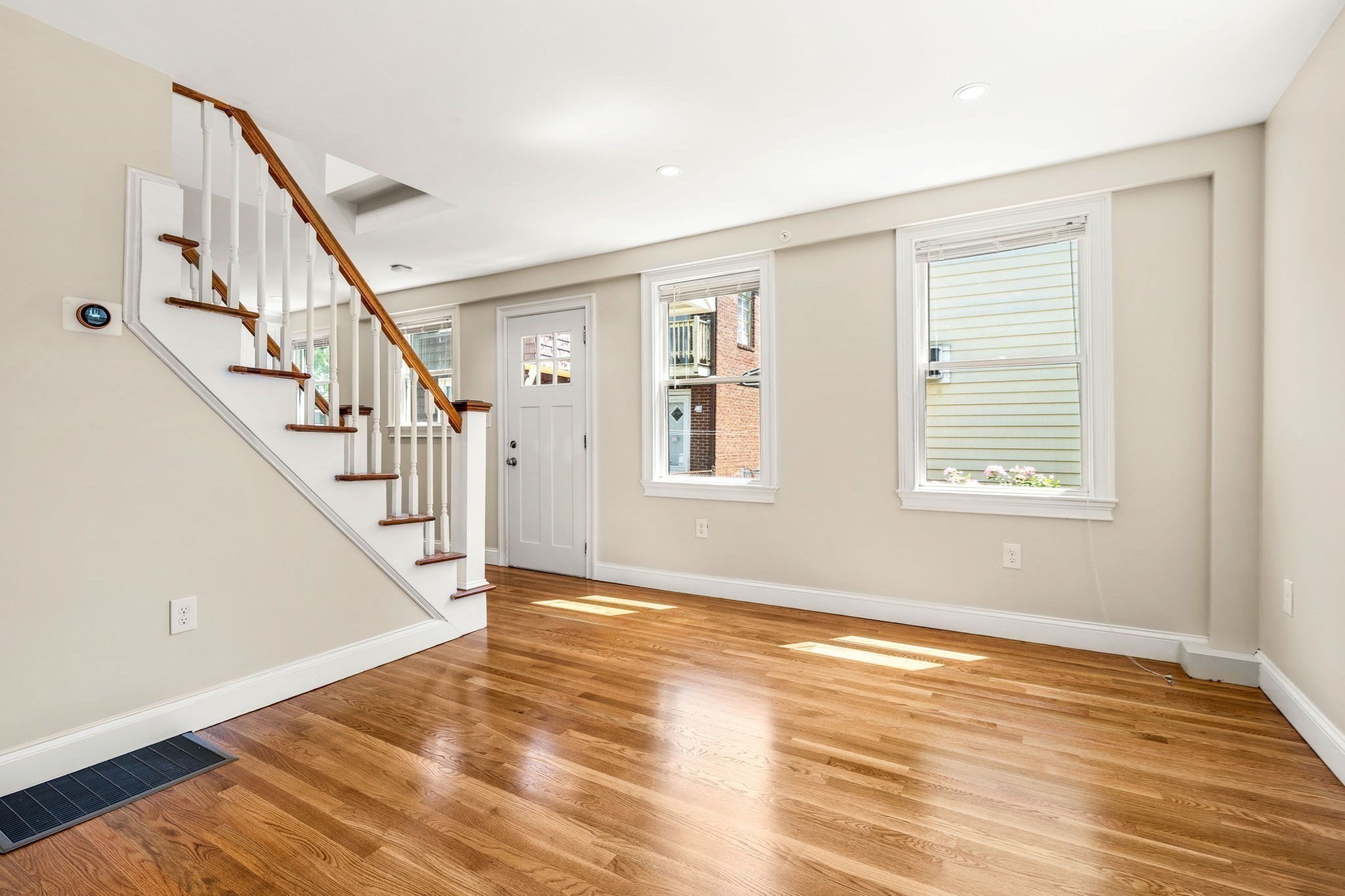 Vacant room with hardwood floors, two windows, and a staircase with a wooden handrail.