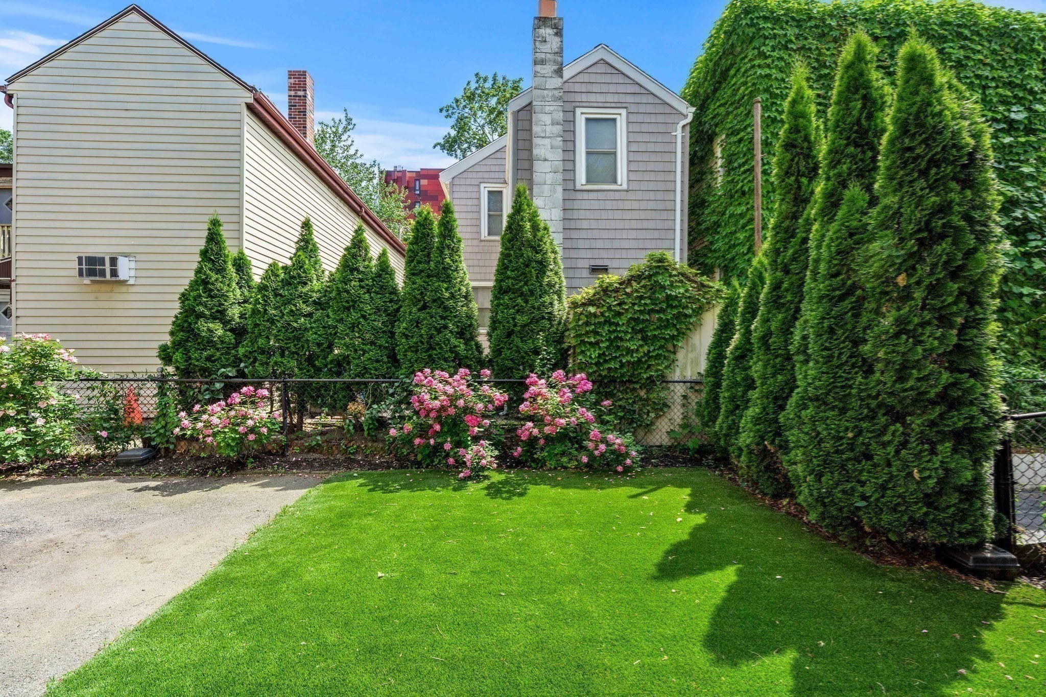 Landscaped yard with a green lawn, pink flowers, tall evergreens, and a gray-shingled house.