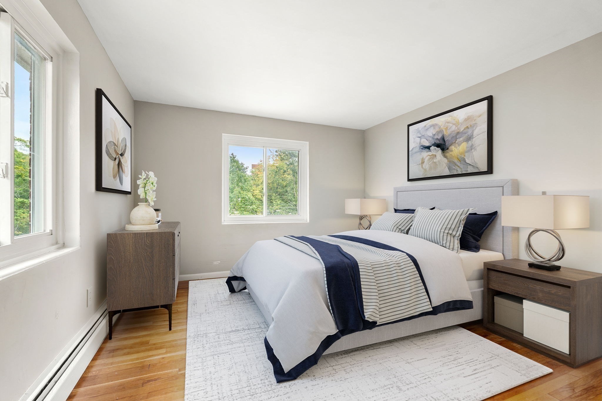 Staged bedroom with a gray upholstered bed, navy-and-white bedding, and windows overlooking trees.