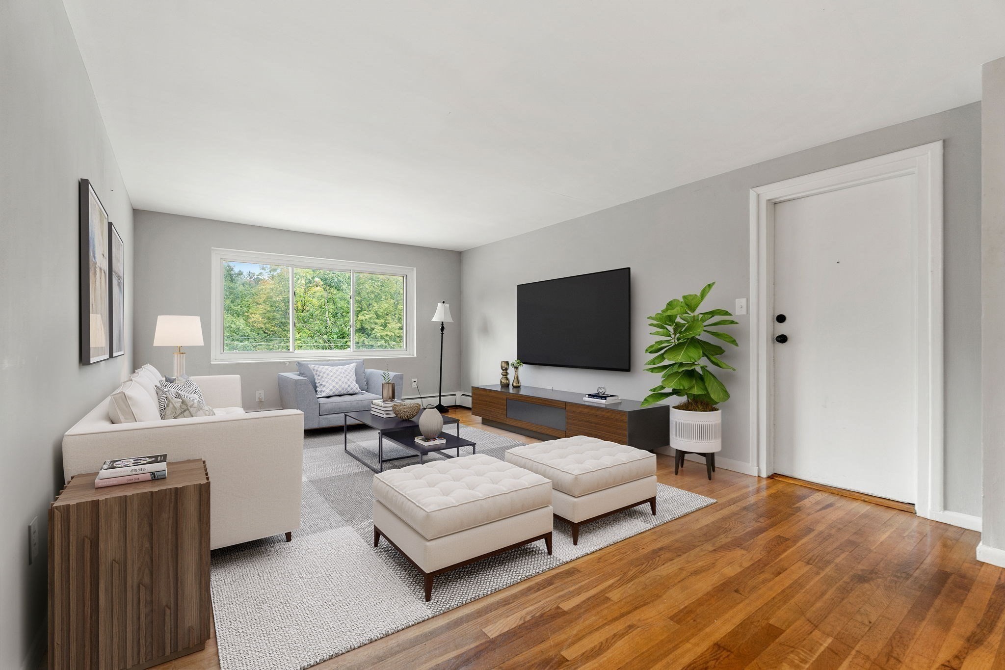 Staged living room with a large window, wall-mounted television, and warm hardwood floors.