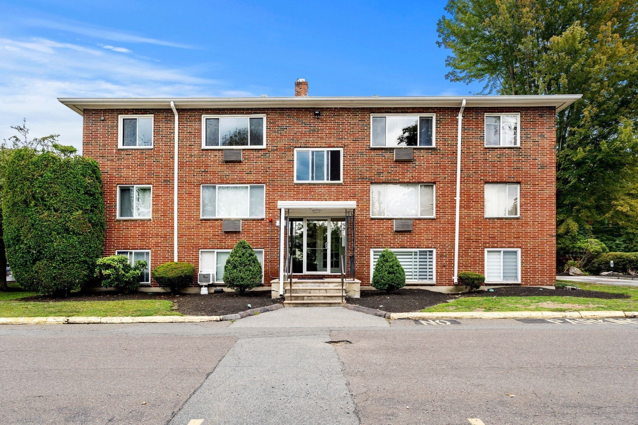 Three-story brick apartment building with a central glass entrance and landscaped shrubs.