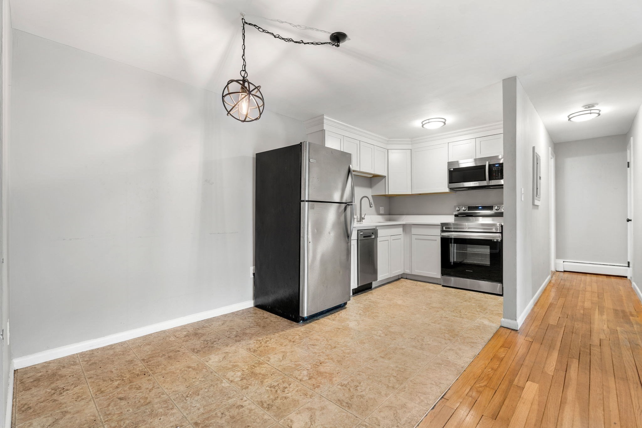 Kitchen with white cabinets, stainless steel appliances, tan tile flooring, and a wood-floored hallway.