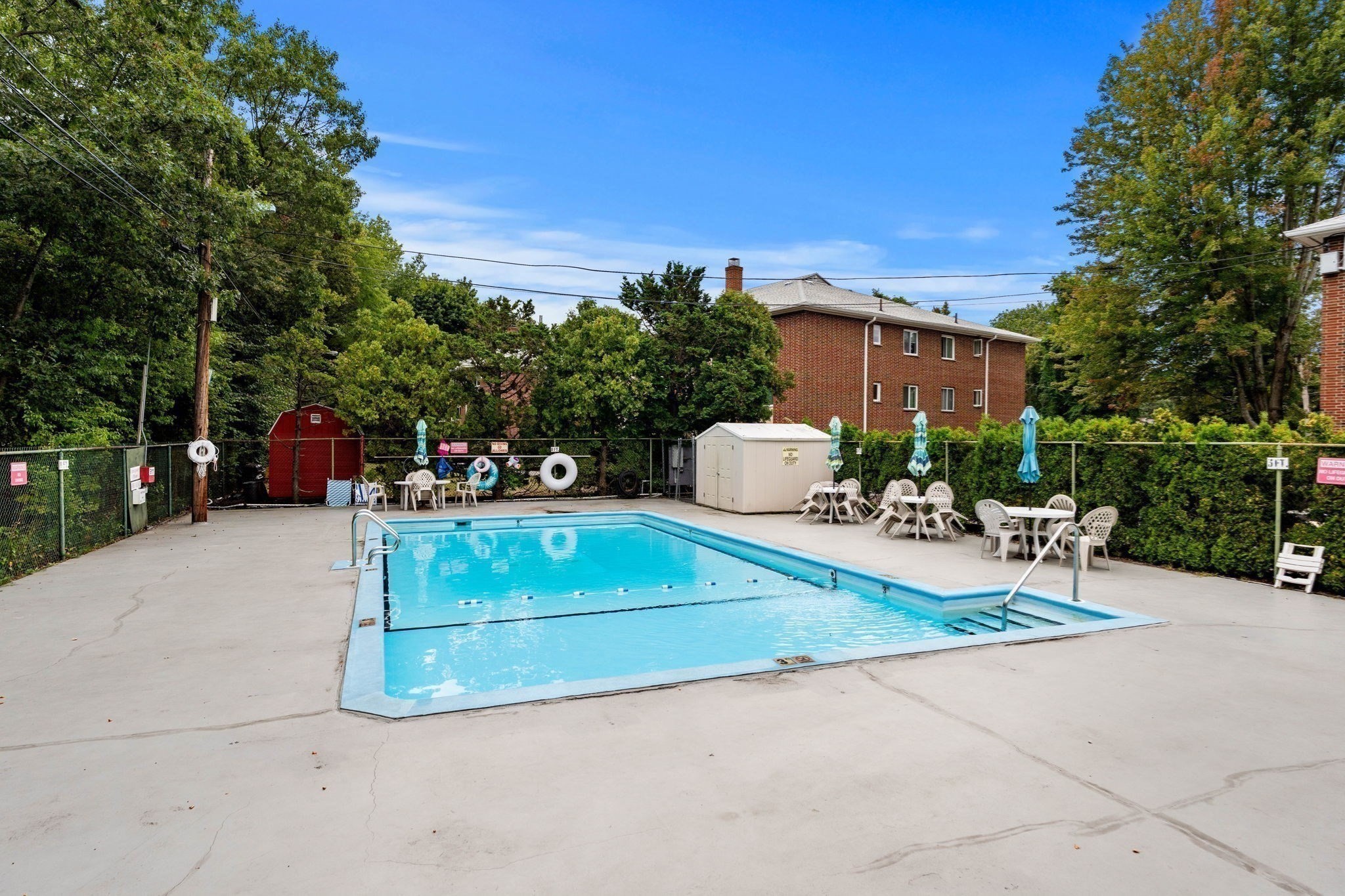 Outdoor pool with a fenced concrete deck, patio seating, umbrellas, and brick apartments behind it.