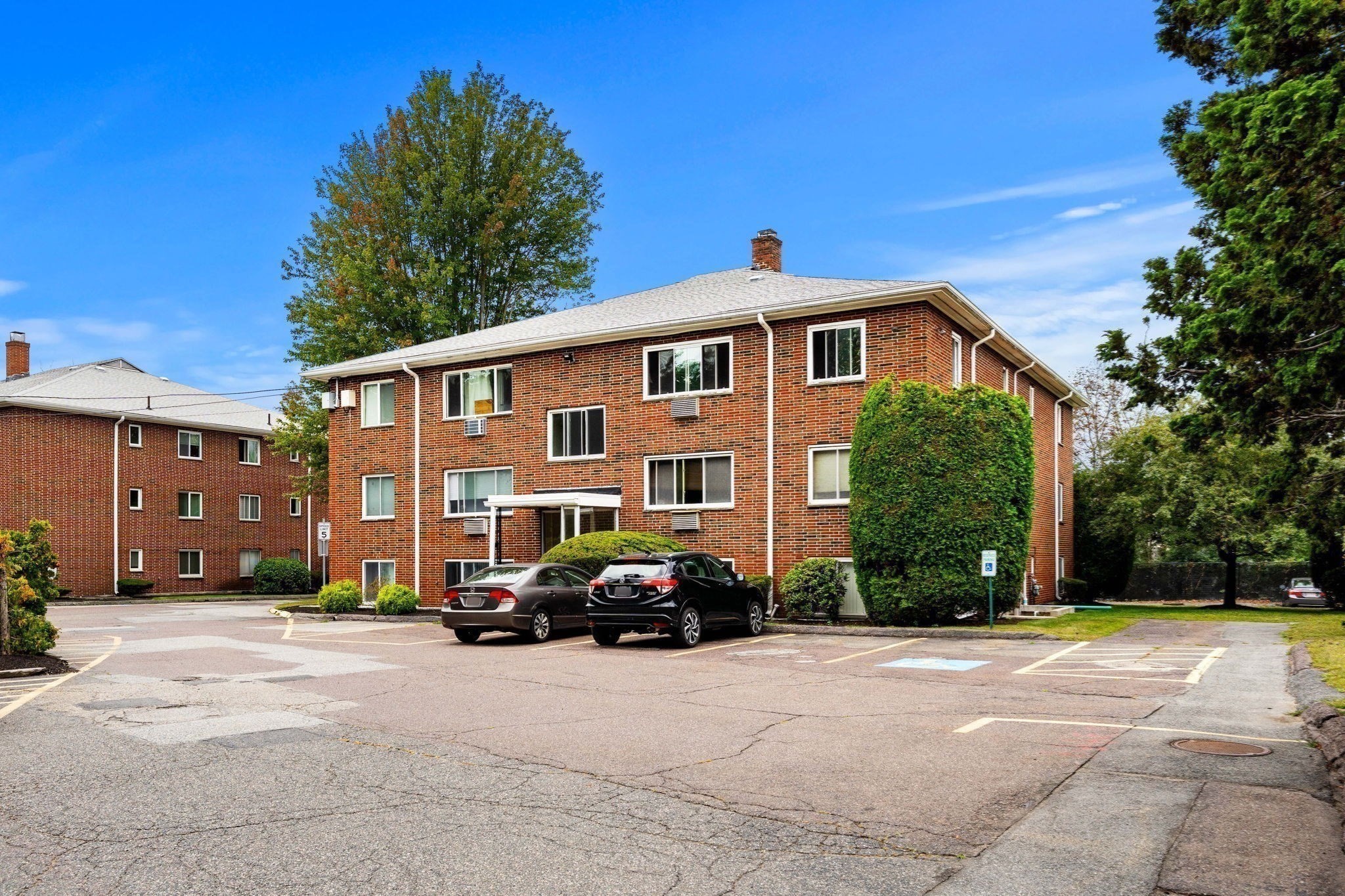 Red-brick condominium building beside a parking lot with marked accessible spaces and parked cars.