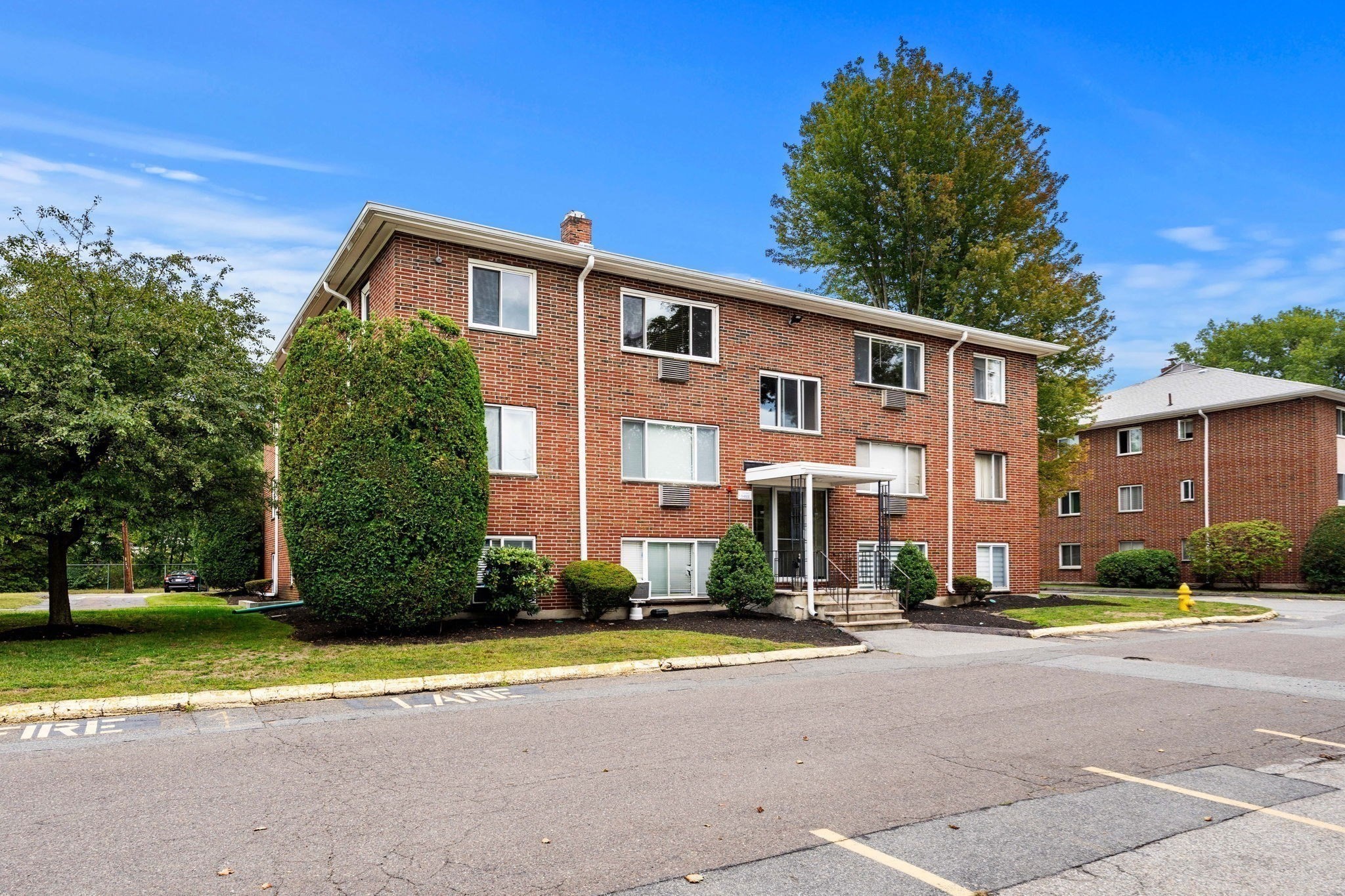 Three-story red-brick apartment building with a covered entrance and landscaped shrubs.