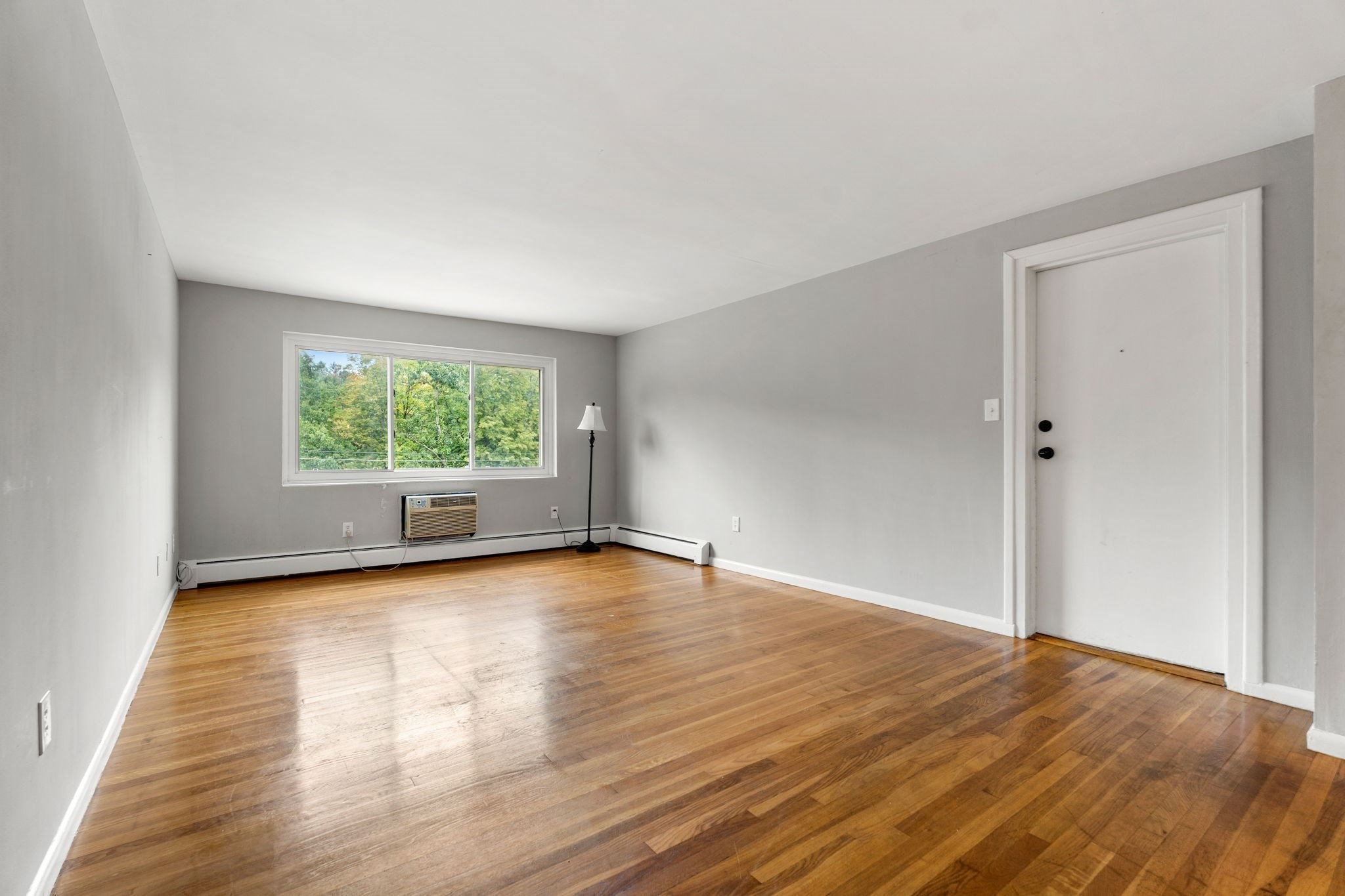 Empty room with polished hardwood floors, a wide window overlooking trees, and a window air conditioner.