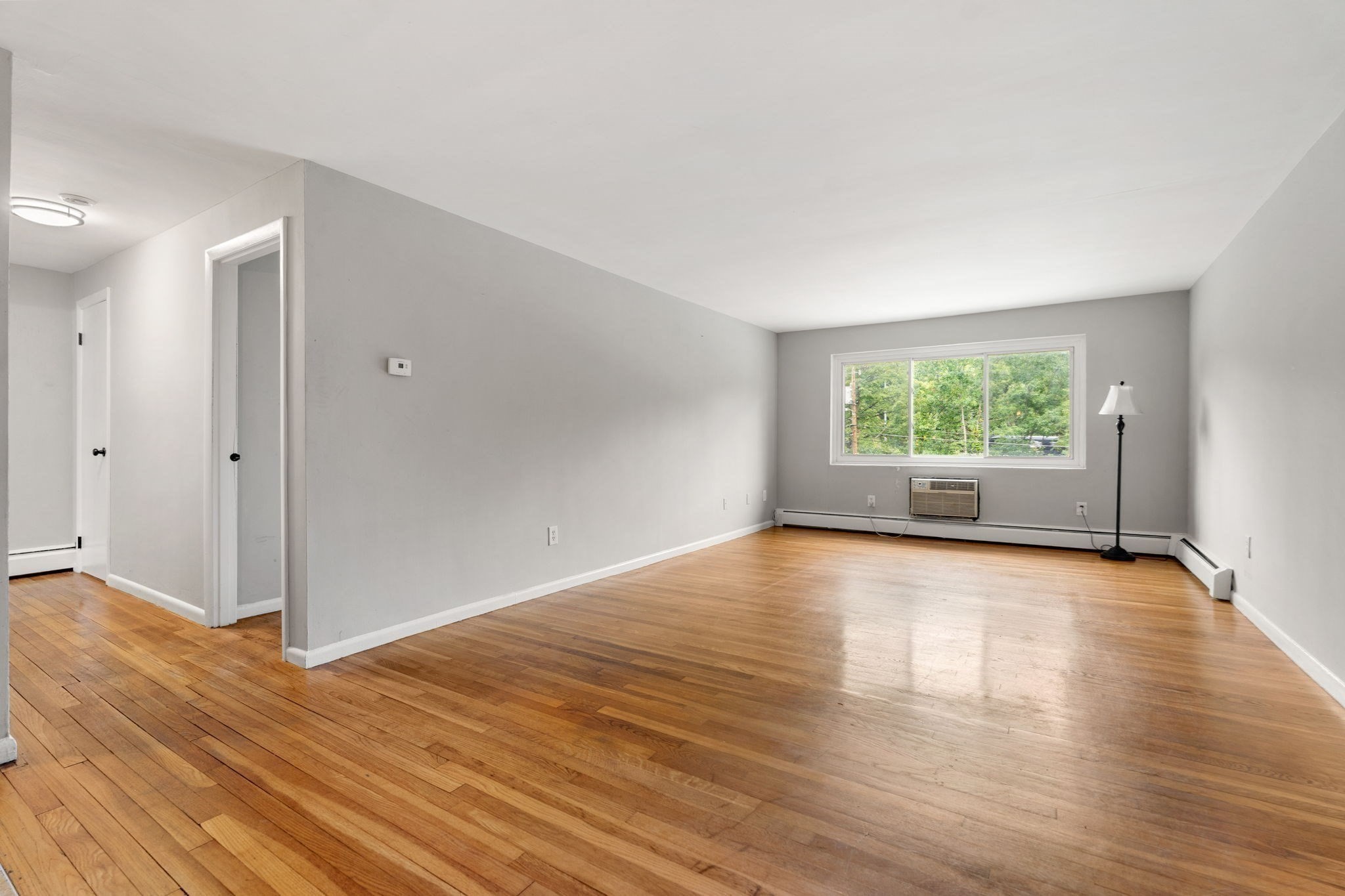 Empty living room with hardwood floors, a wide window, and a wall-mounted air conditioner.