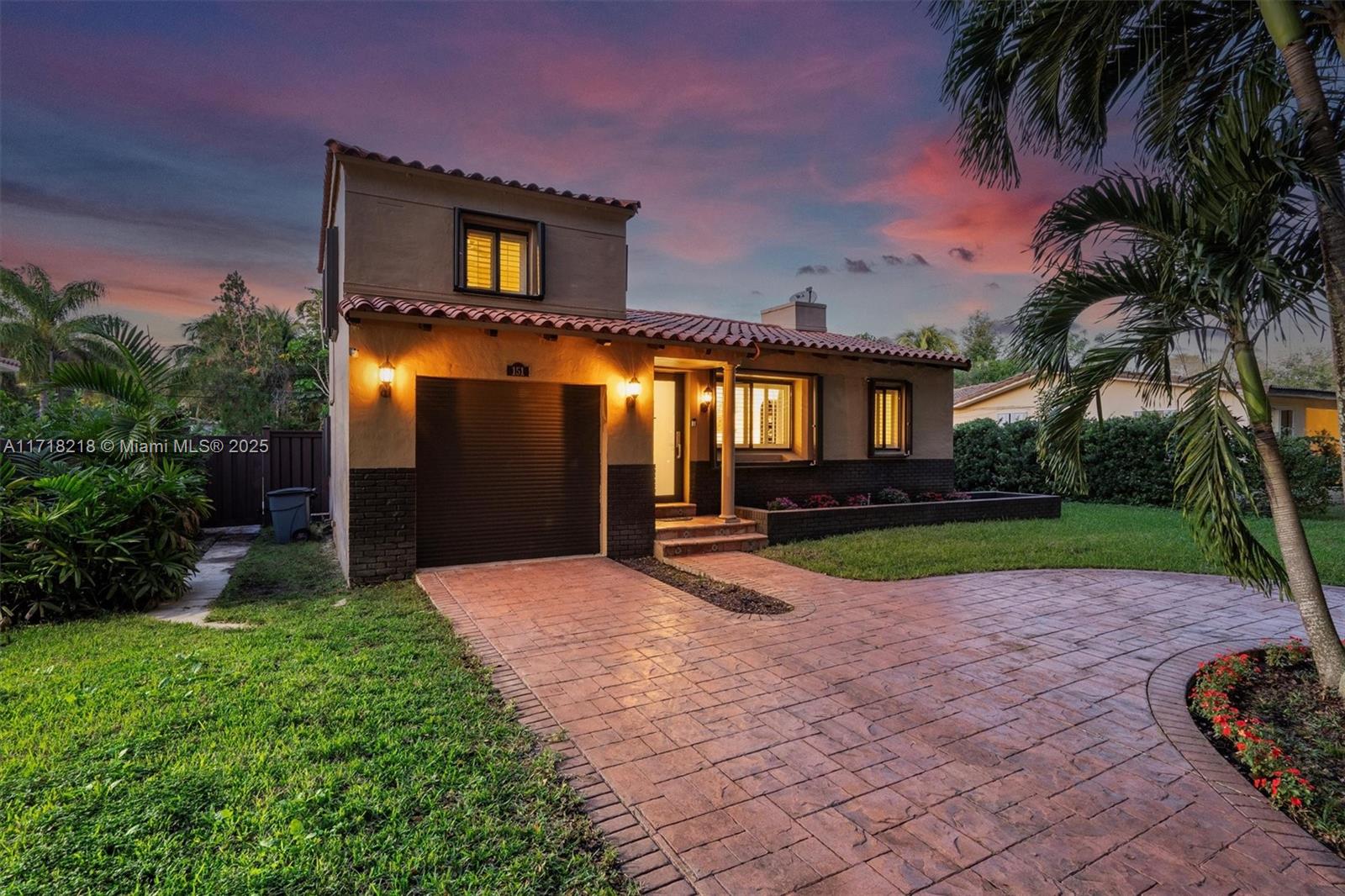 Two-story stucco house with an attached garage and brick-paved driveway beneath a colorful dusk sky.