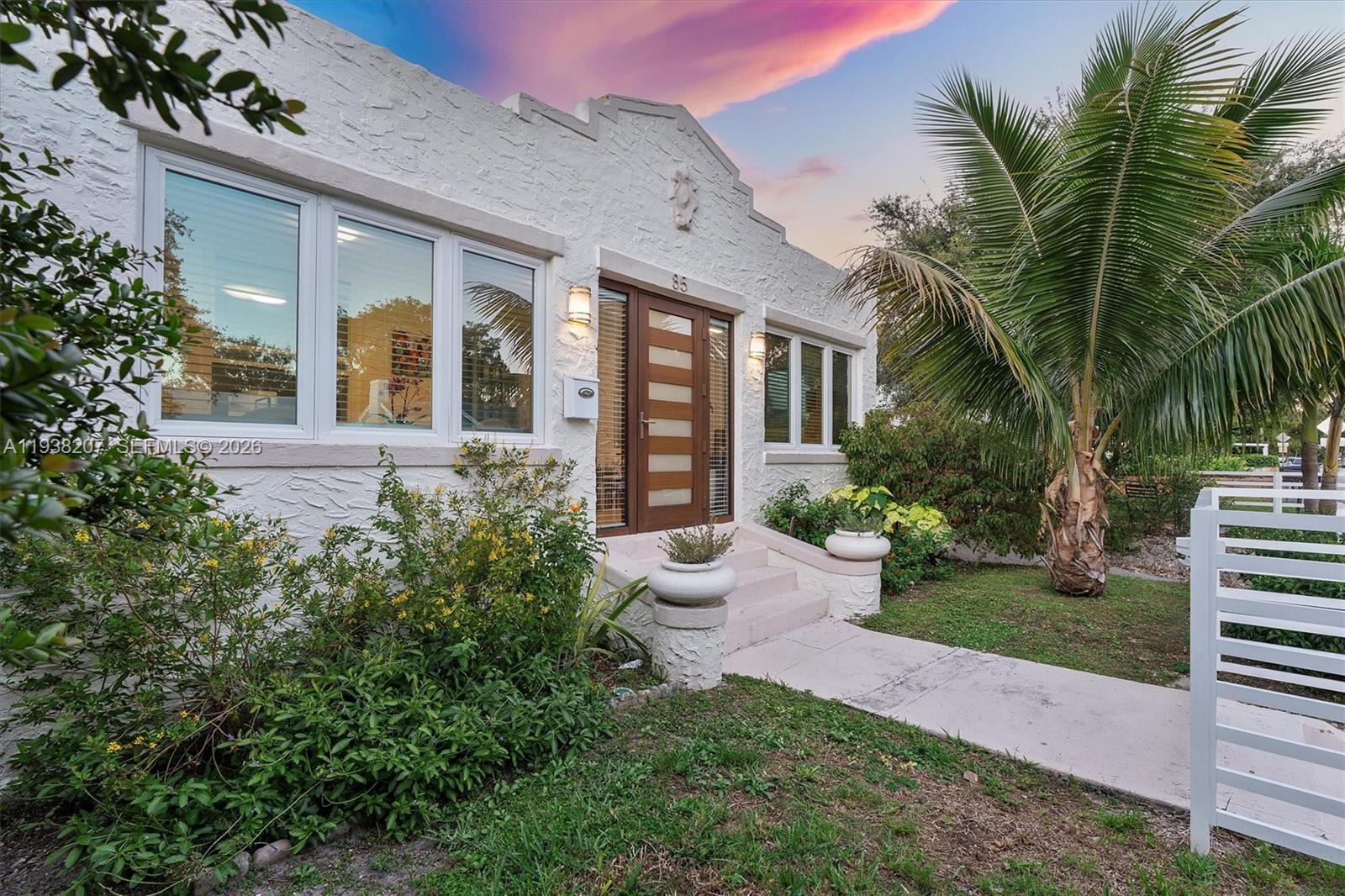White stucco home with a modern wood entry door, tropical palms, and a pink dusk sky.